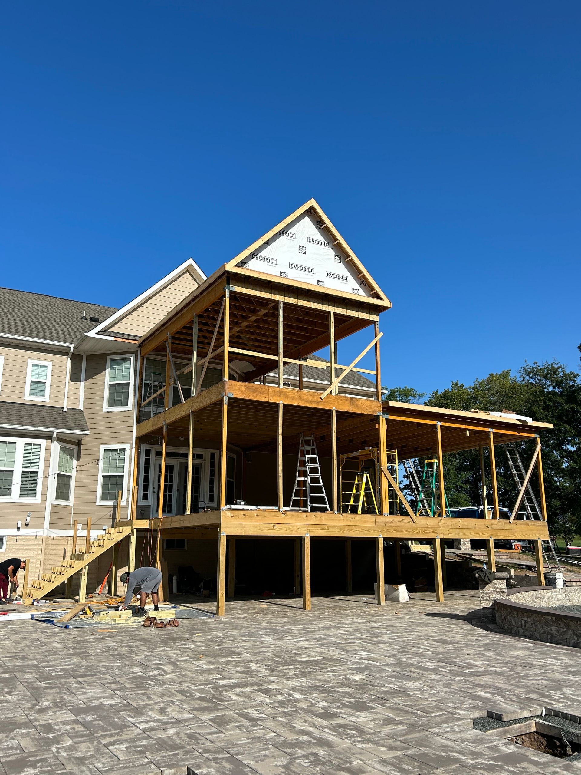 Two-story wooden deck under construction on the back of a house; workers, tools, and materials visible.