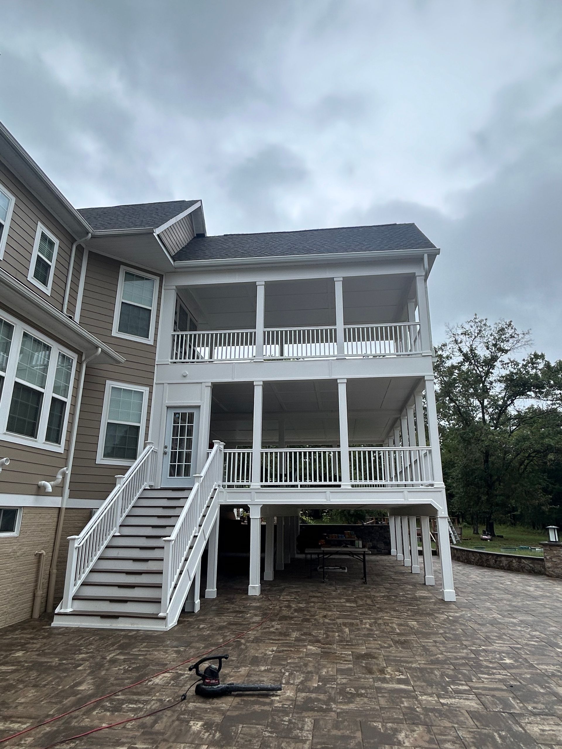 Two-story white screened porch with stairs. Beige house on left, cloudy sky. Brick ground, lawn equipment in the foreground.