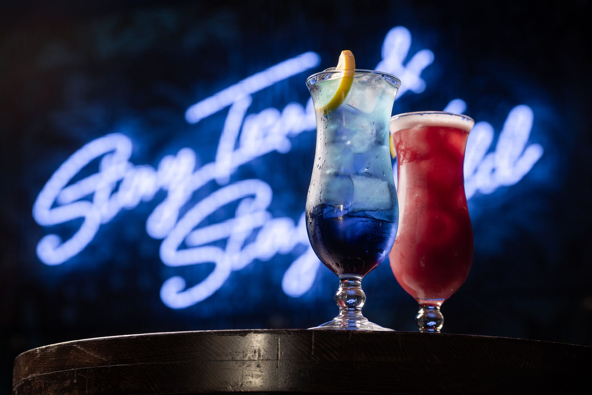 Two cocktails are on a table in front of a neon sign that says stay tonight