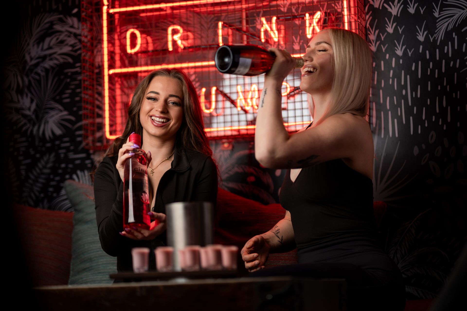 Two women are sitting at a table drinking wine in front of a neon sign.