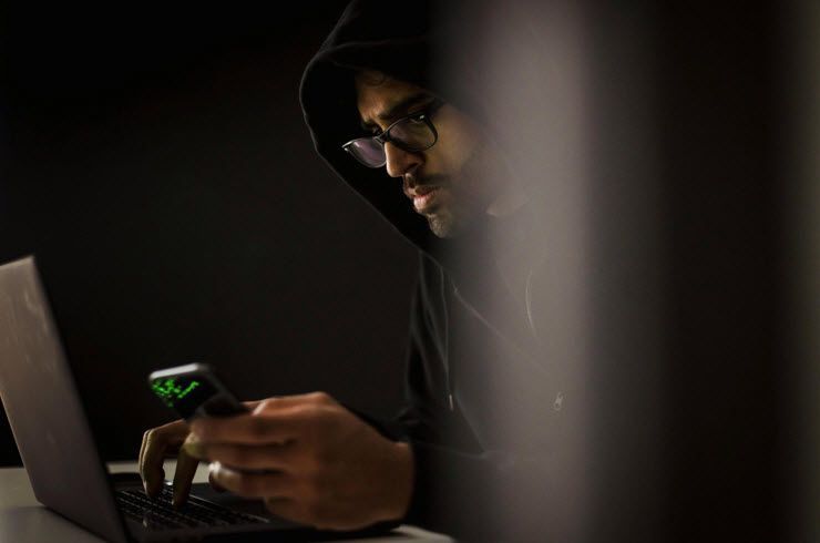 A man in a hood is using a laptop and a cell phone.