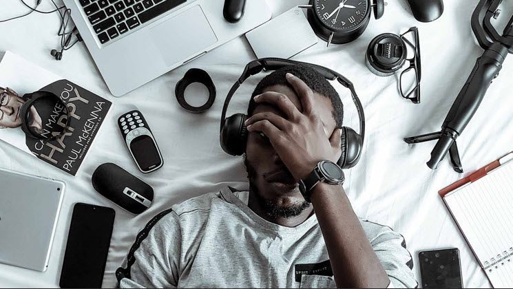 A man wearing headphones is laying on a bed surrounded by electronics.