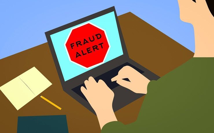 A person is typing on a laptop with a fraud alert sign on the screen.