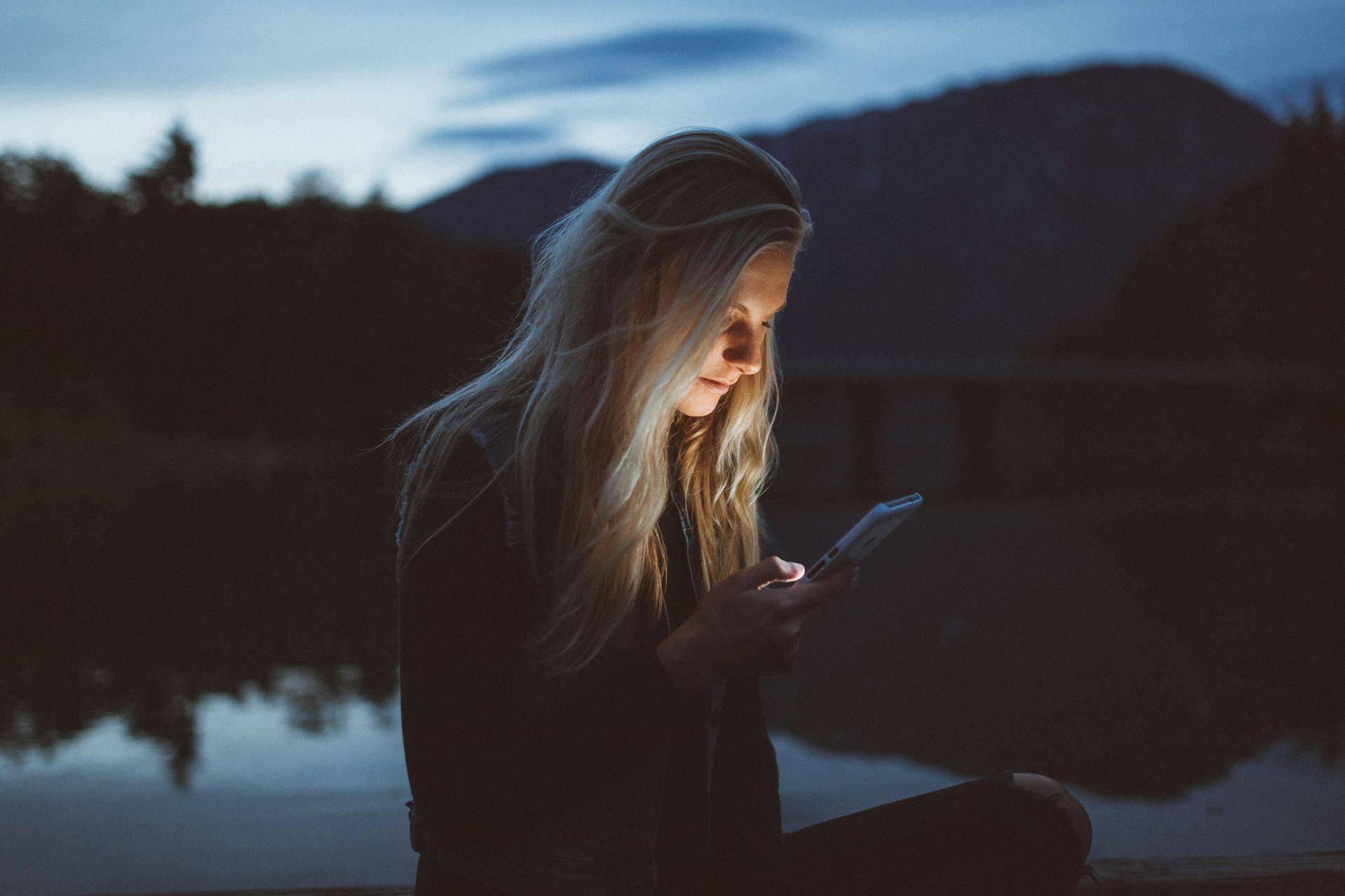 A woman is sitting by a lake at night looking at her cell phone.