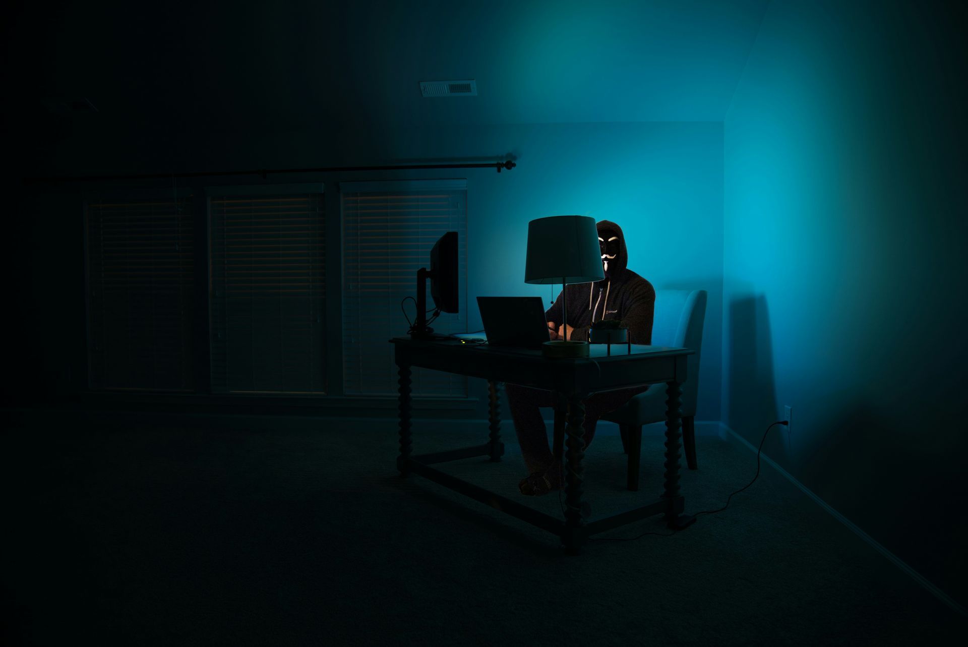 A man in a mask is sitting at a desk in front of a computer in a dark room.