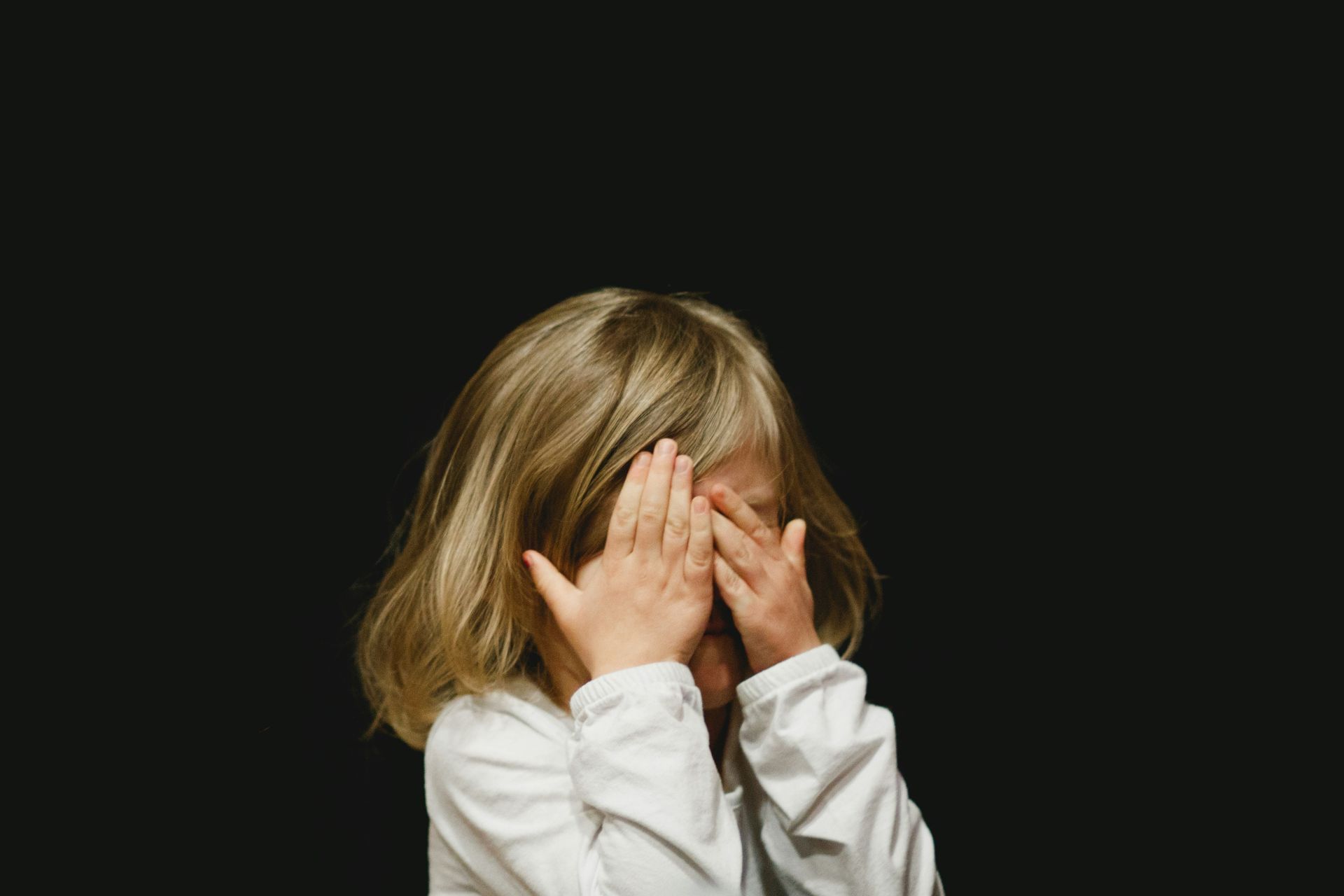 A little girl is covering her face with her hands on a black background.