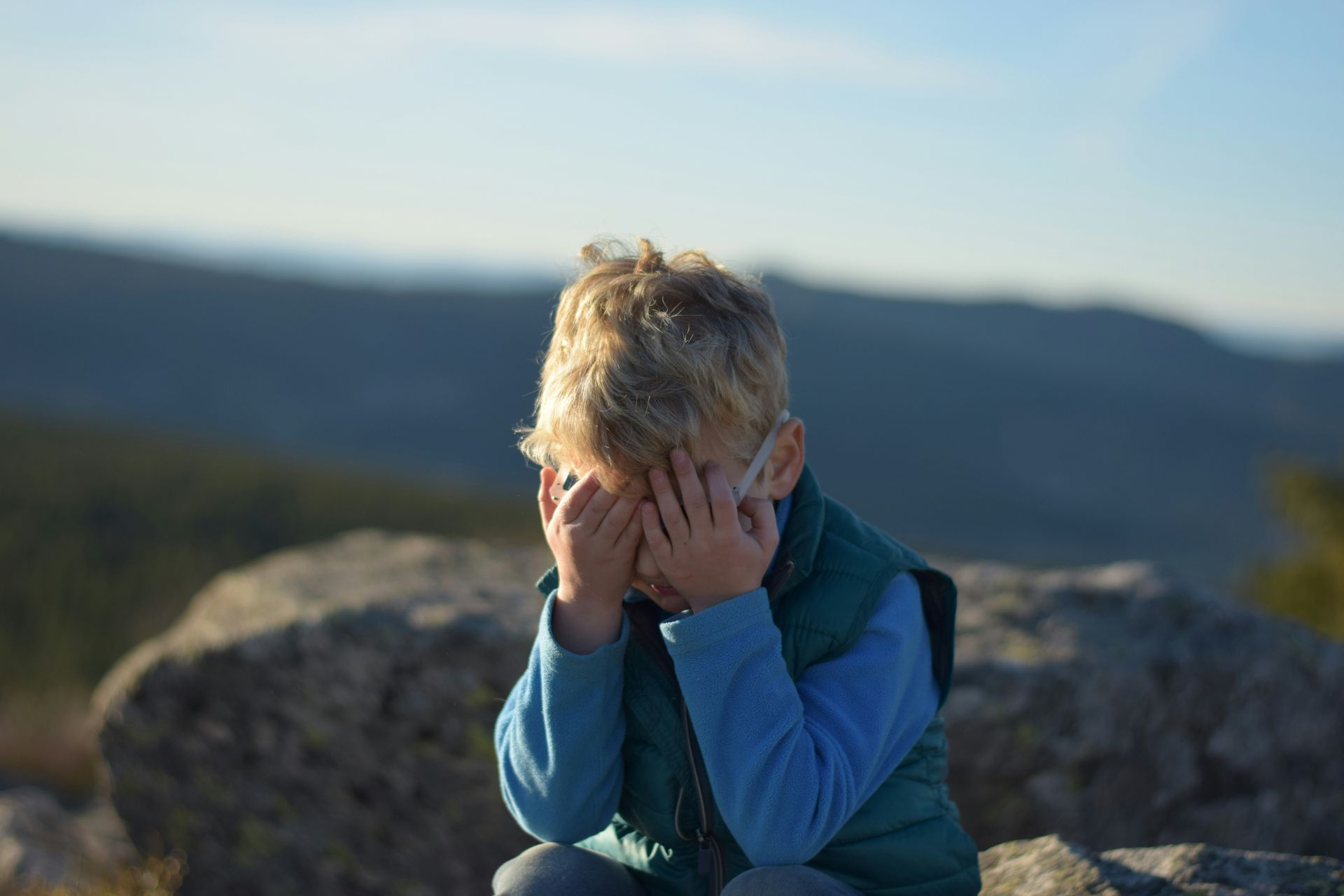 A young boy is sitting on a rock covering his face with his hands.