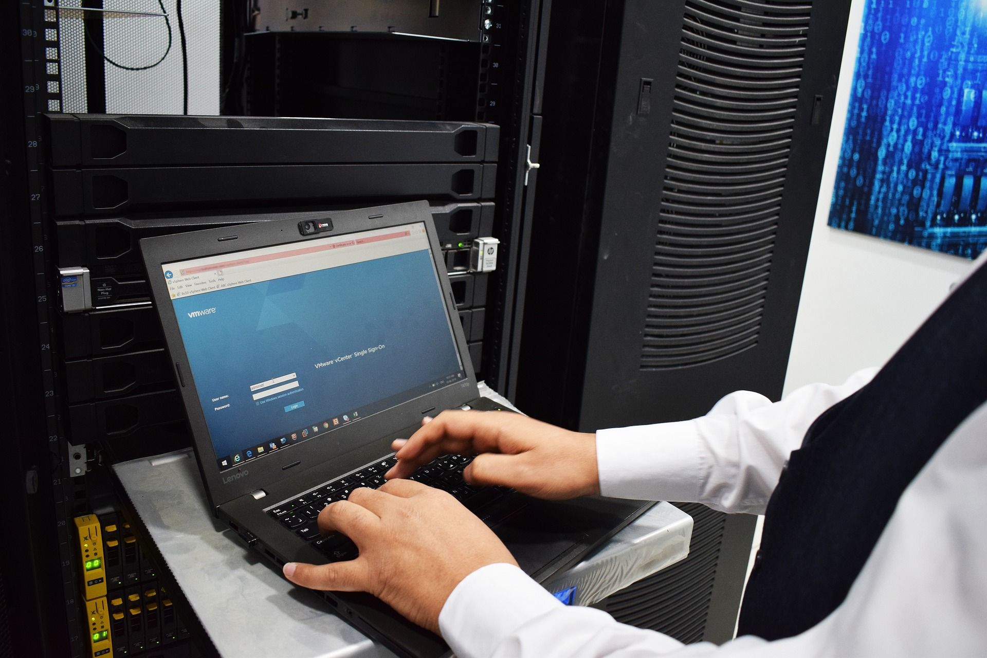 A man is typing on a laptop computer in a server room.