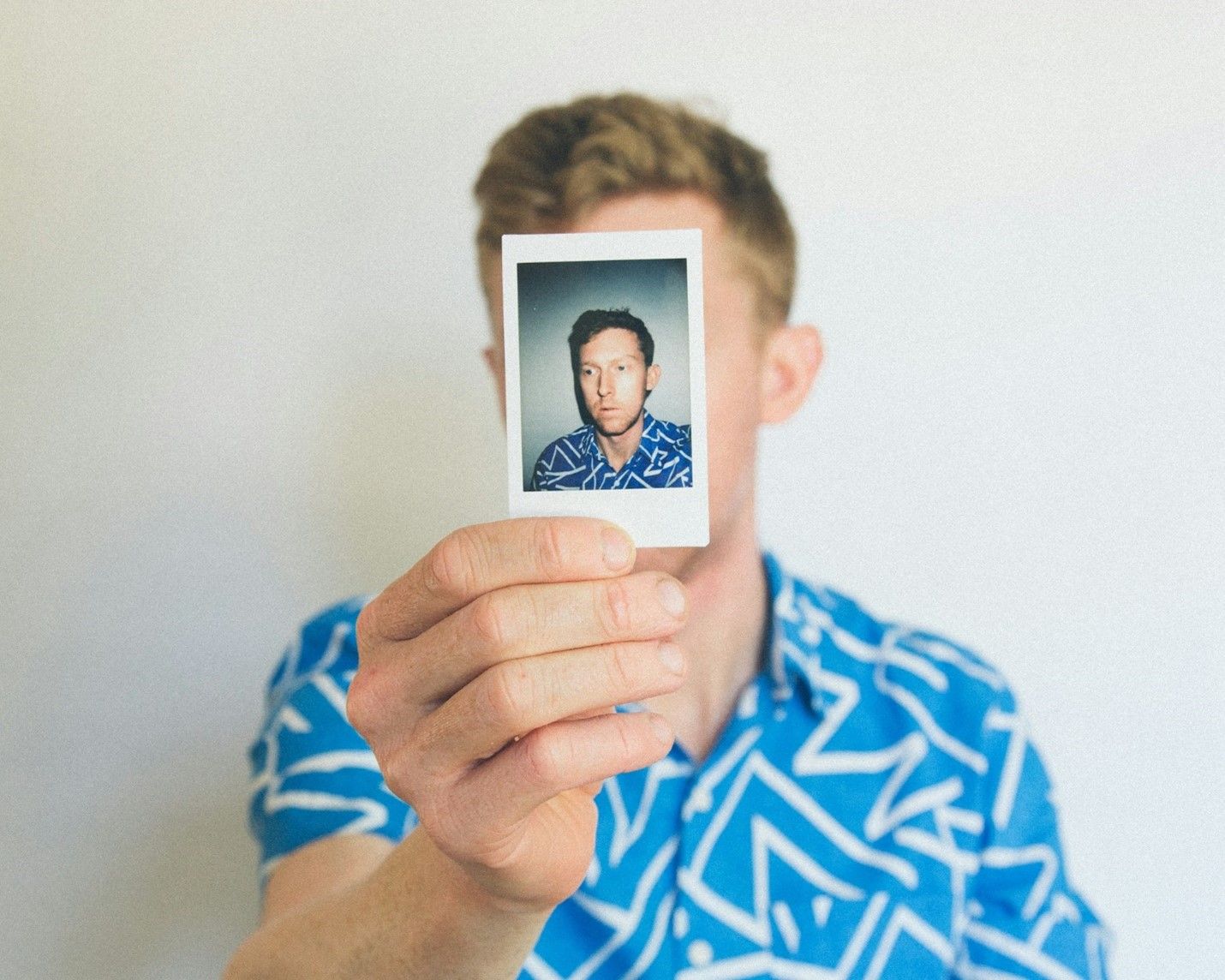 A man in a blue shirt is holding a polaroid picture in front of his face.