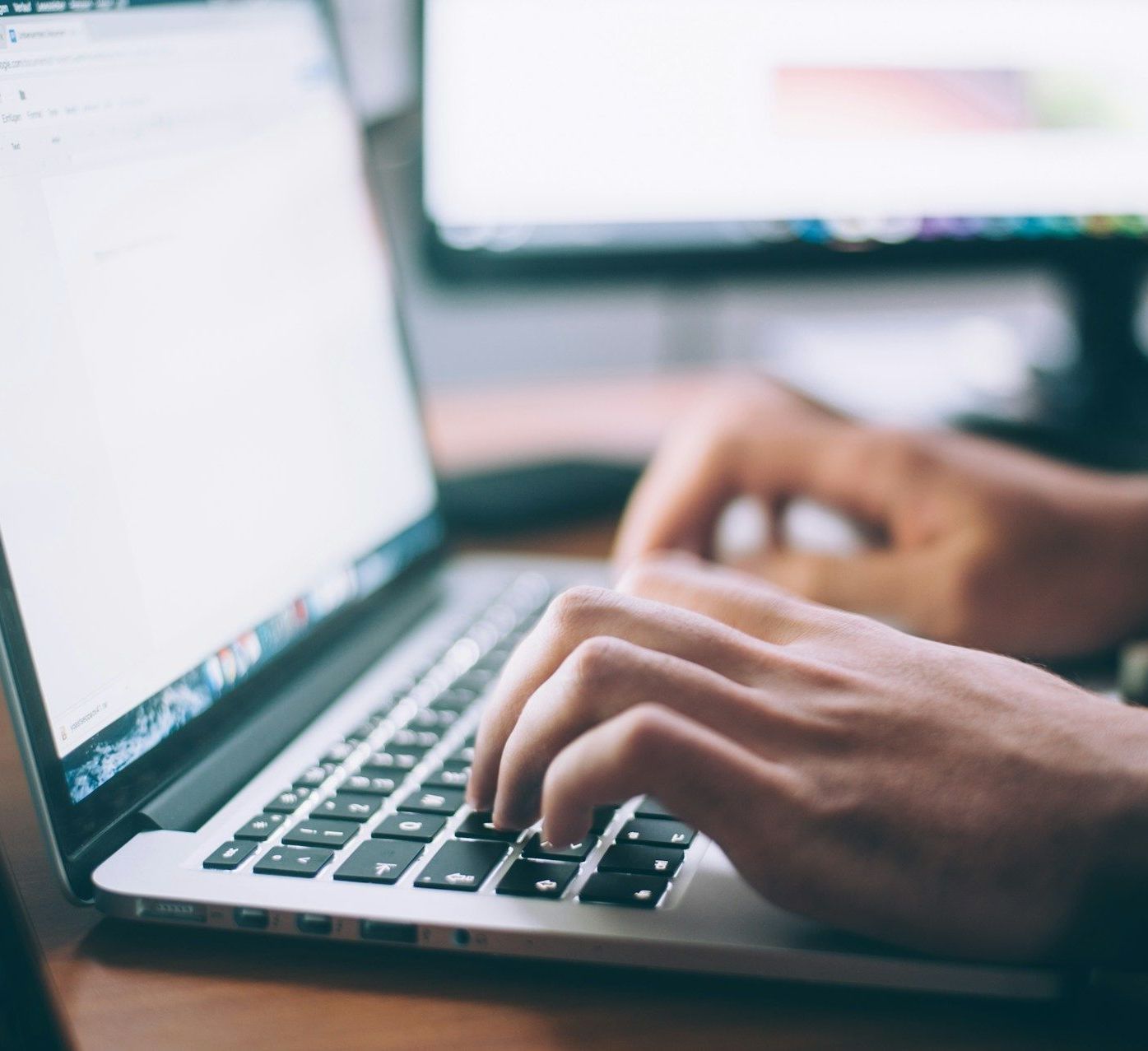 A person is typing on a laptop with a monitor in the background