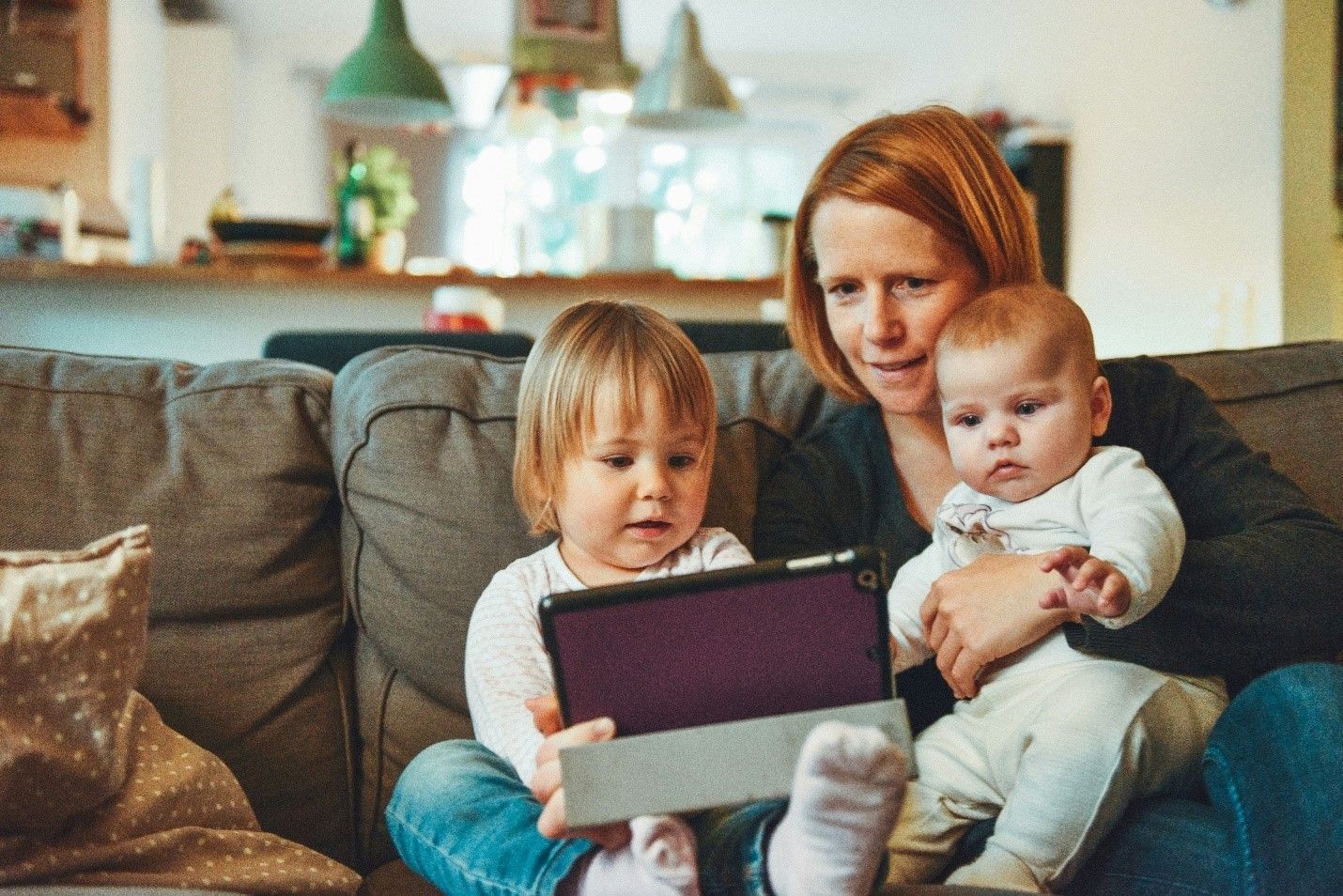 A woman is sitting on a couch holding two children and looking at a tablet.