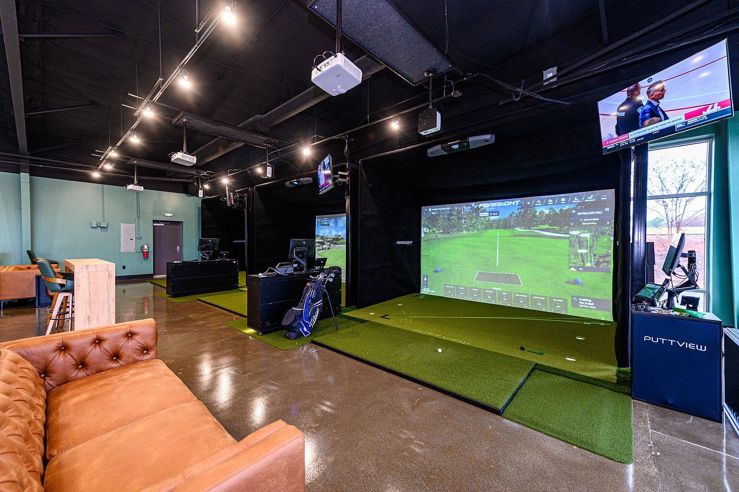 Indoor golf simulator with green turf, screen displaying a golf course, and a concrete wall.