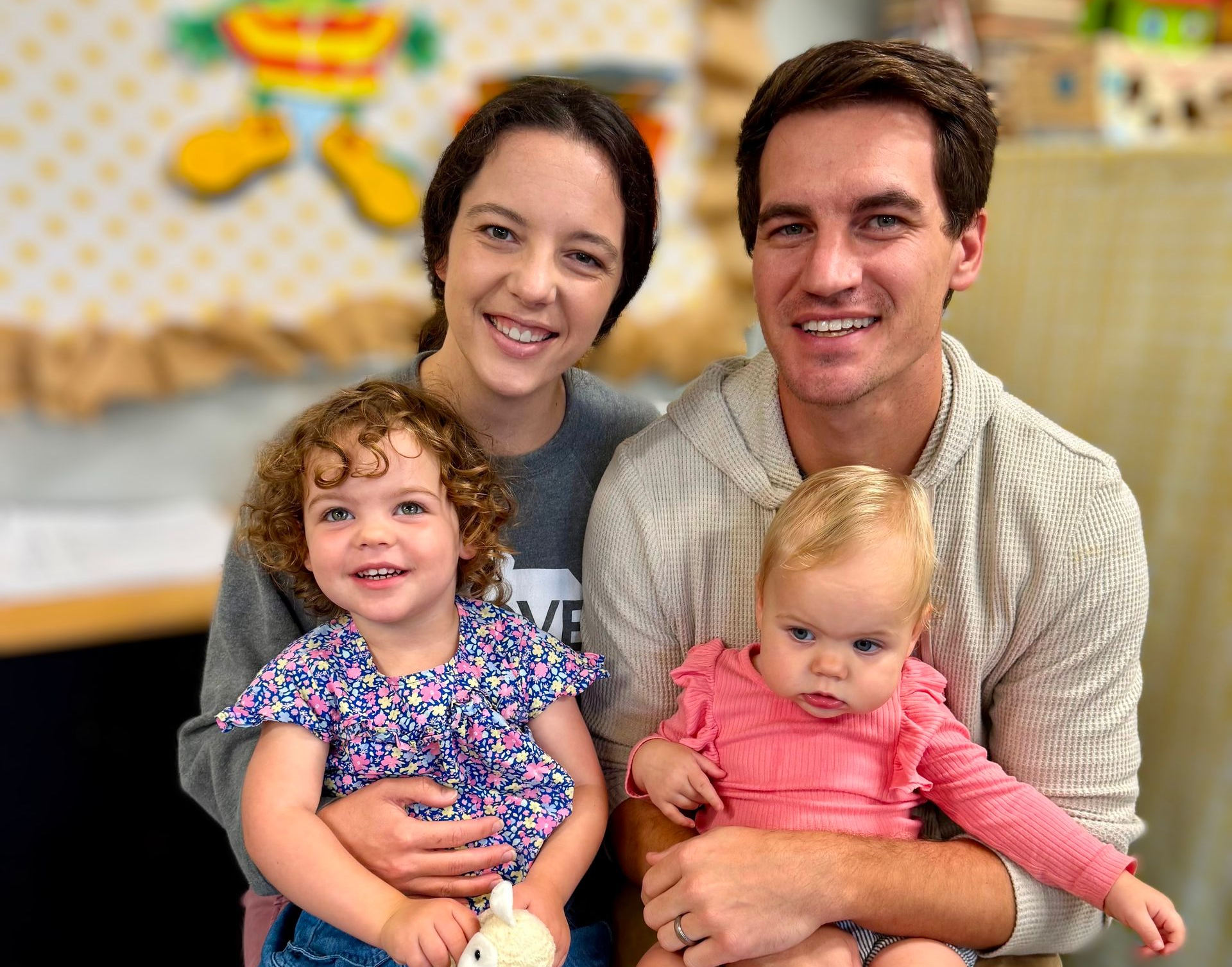 Family of four smiling, sitting indoors; two young children, parents.