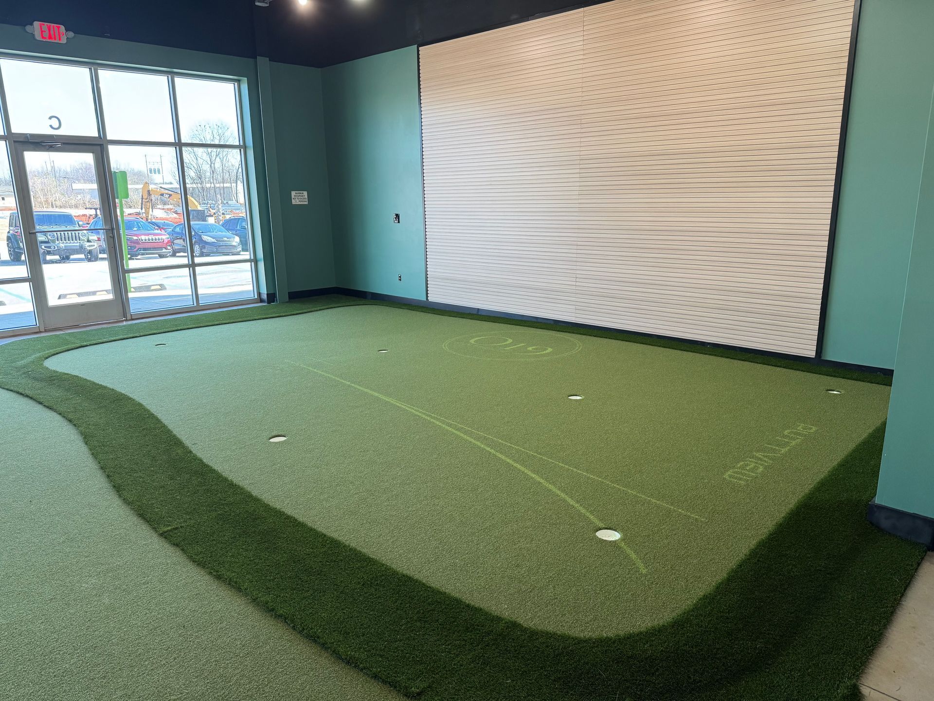 Man swings golf club on a green indoor putting area with white target markers. Black wall with text.