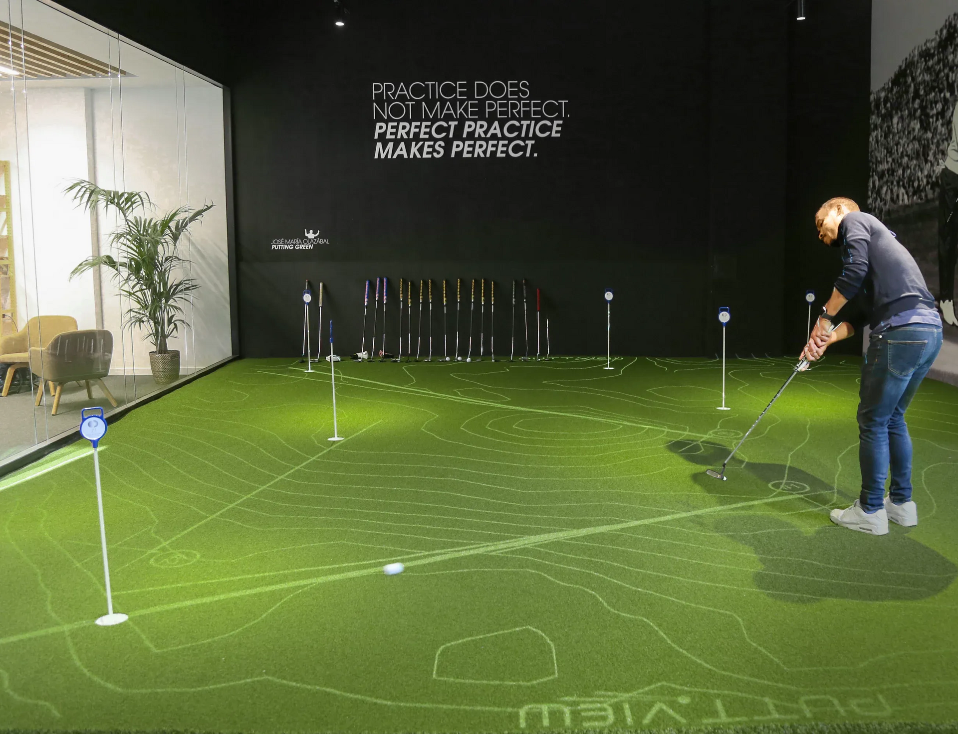 Man swings golf club on a green indoor putting area with white target markers. Black wall with text.