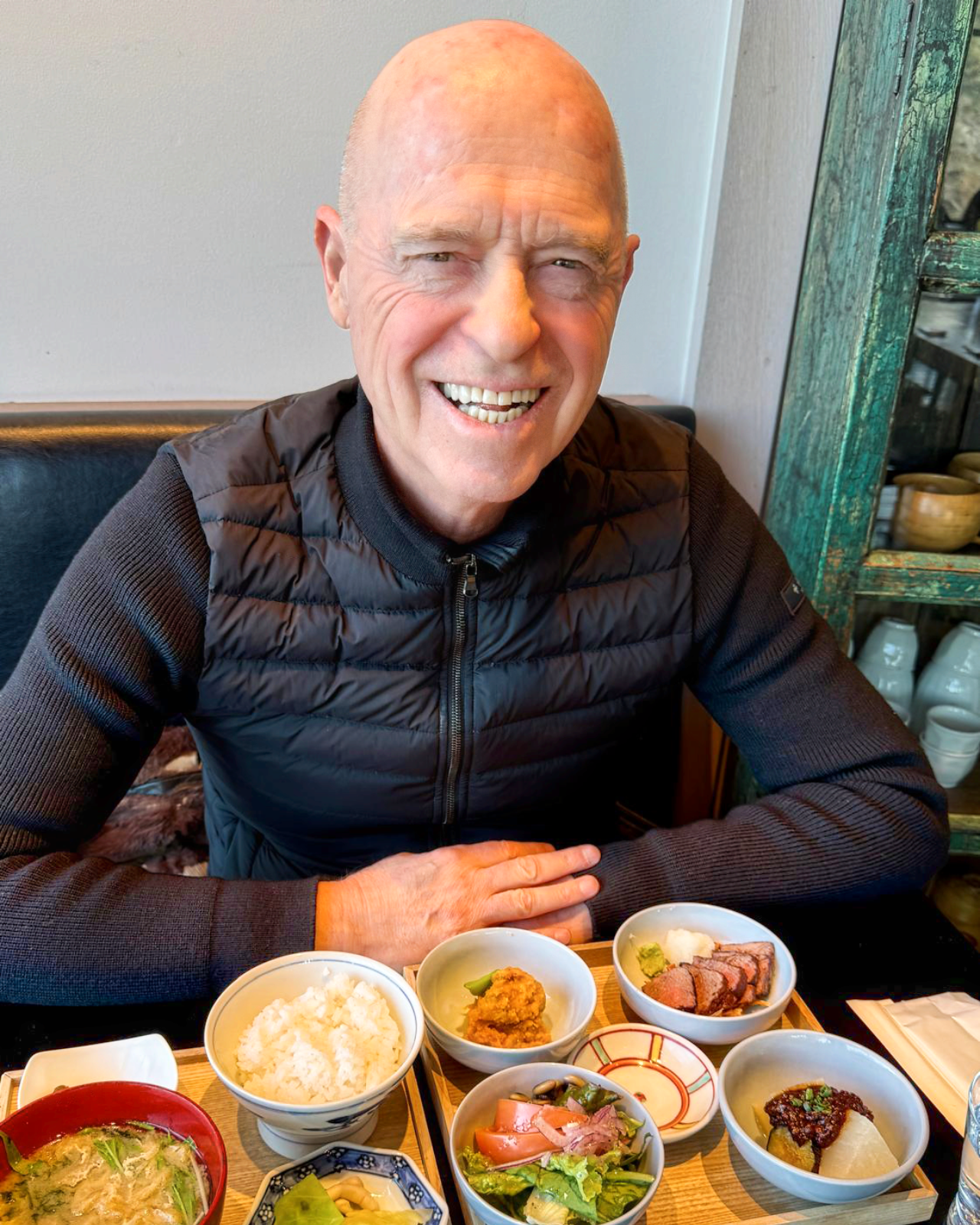 Bald man smiles at the camera, seated at a table with a Japanese meal, black vest, light background.