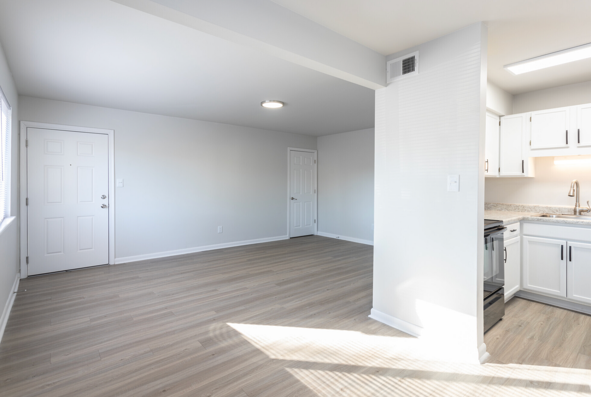 An empty living room with hardwood floors and a kitchen in the background.