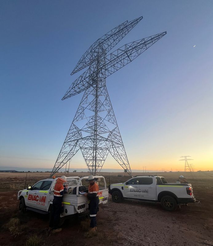 Two workers next to white trucks with logos, in front of a tall metal power pylon at dawn.