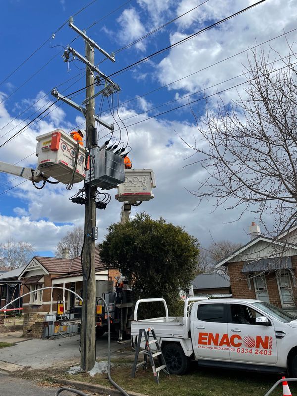 Linemen working on electrical equipment on a utility pole. White bucket trucks, houses, and a blue sky.
