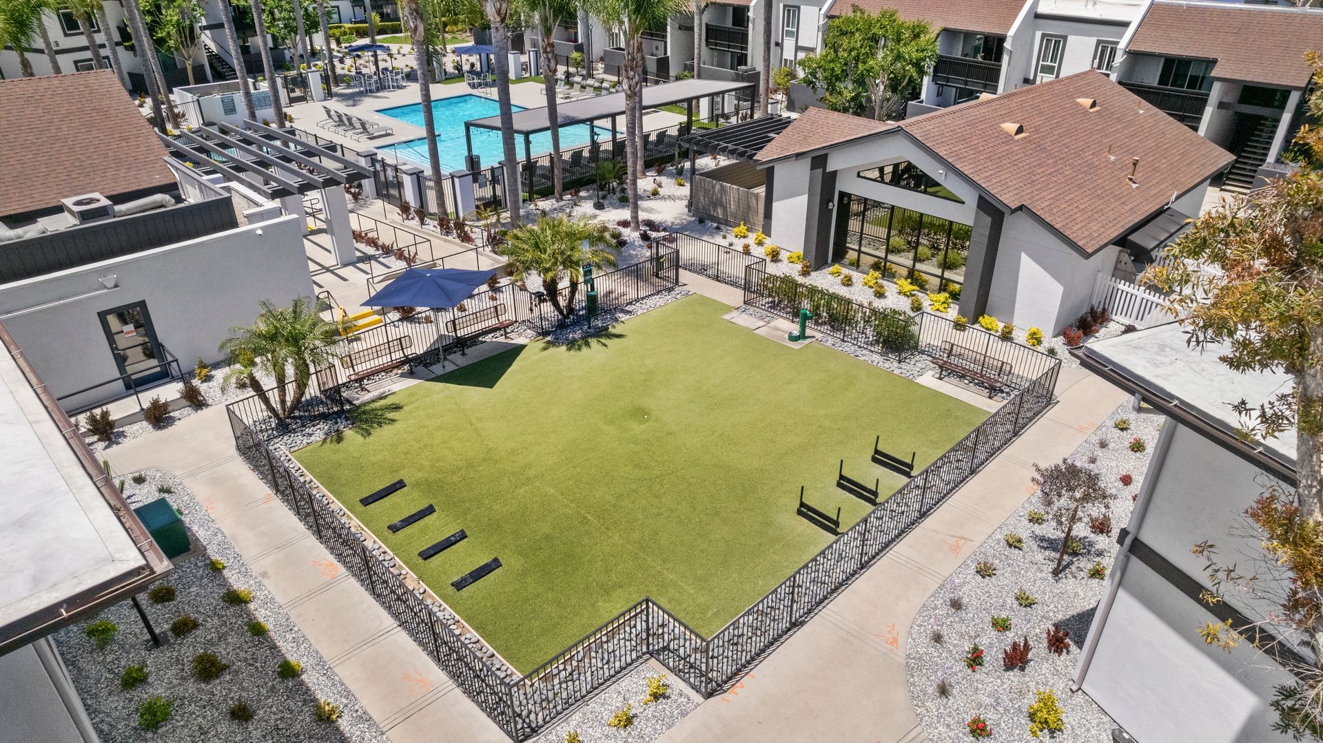 An aerial view of a large grassy area dog park surrounded by buildings and a pool.