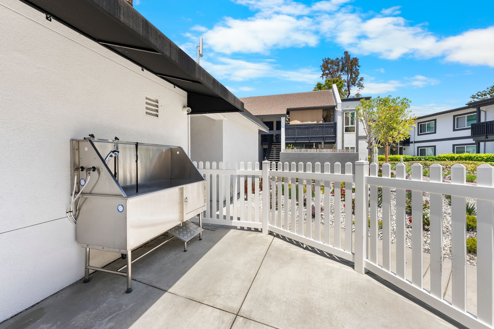 A stainless steel dog wash tub is sitting outside of a building next to a white fence.