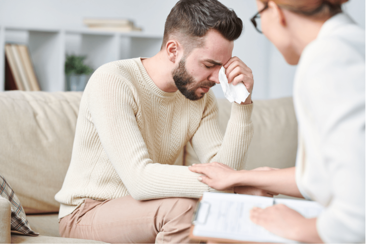 A man is crying while sitting on a couch while a woman comforts him.