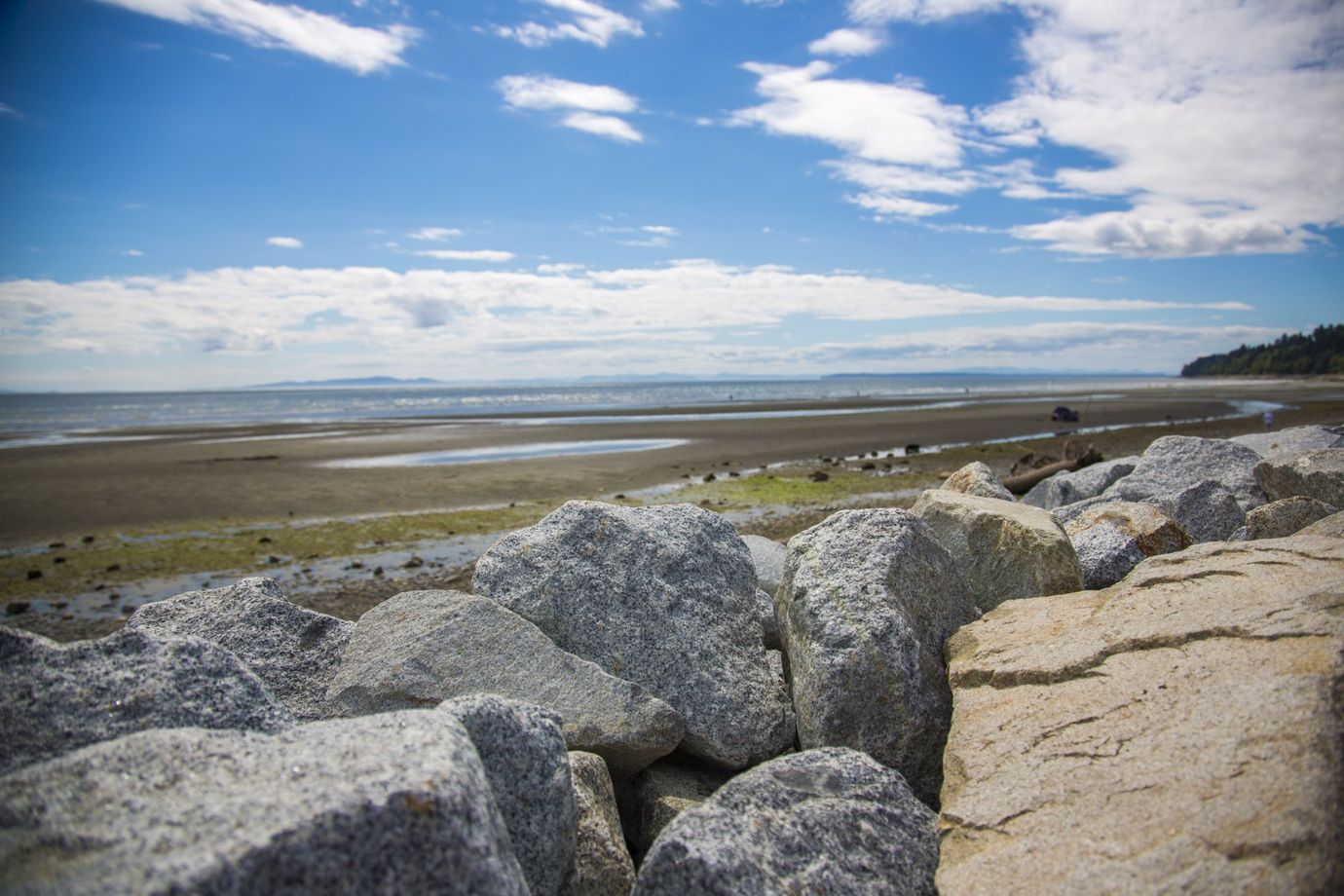 Rocky shoreline with a sandy beach, trees, and blue sky with clouds.