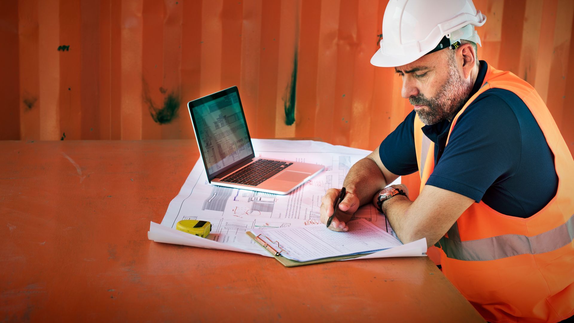 A construction worker is sitting at a table with a laptop and blueprints.