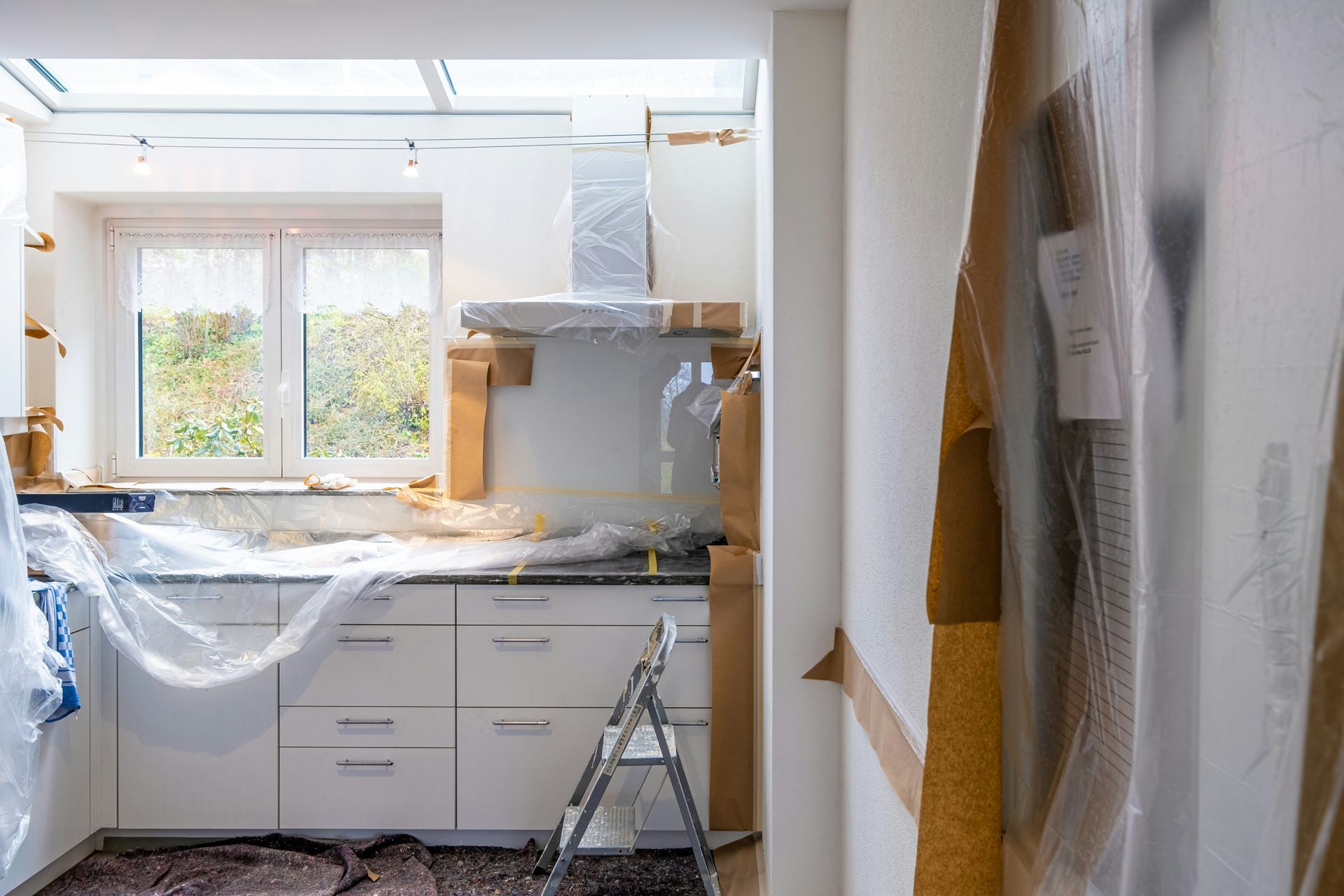 Kitchen undergoing renovation, with white cabinets, countertops covered in plastic, and a ladder.