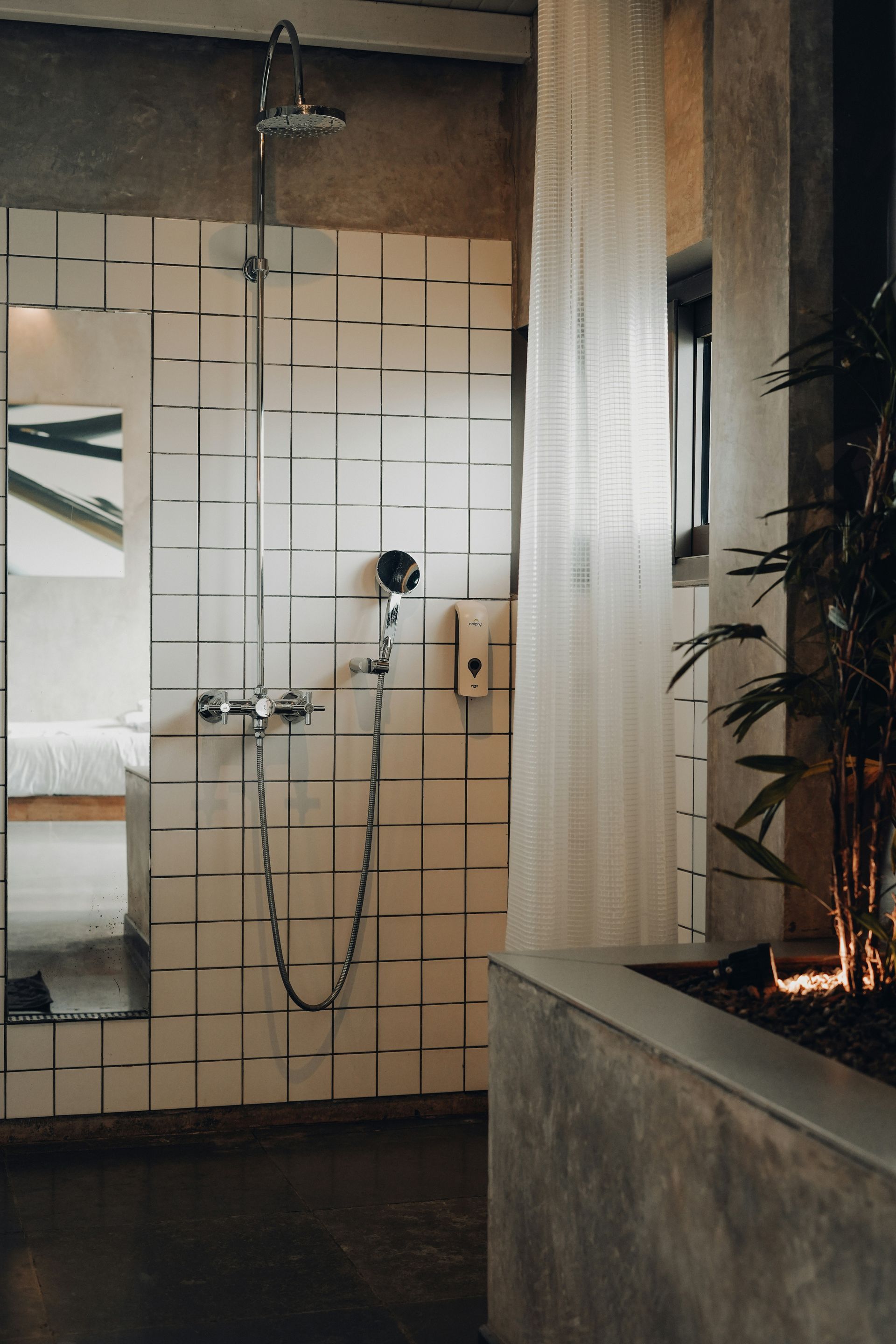 A dim bathroom with white tiles, a tall mirror, sheer white curtain and plants.