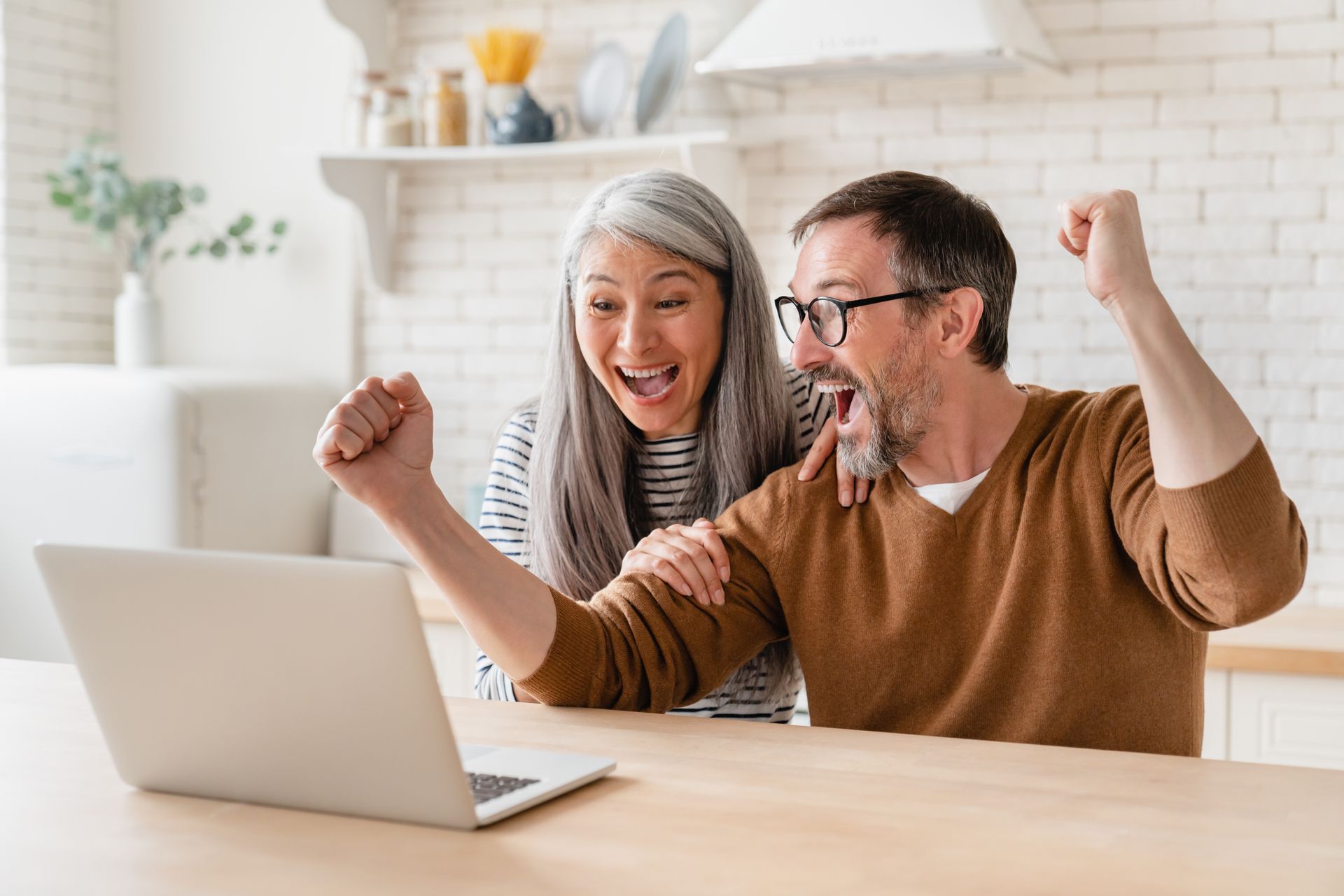 Excited older couple cheering, looking at laptop in a kitchen setting. Man raises fist, woman smiles.