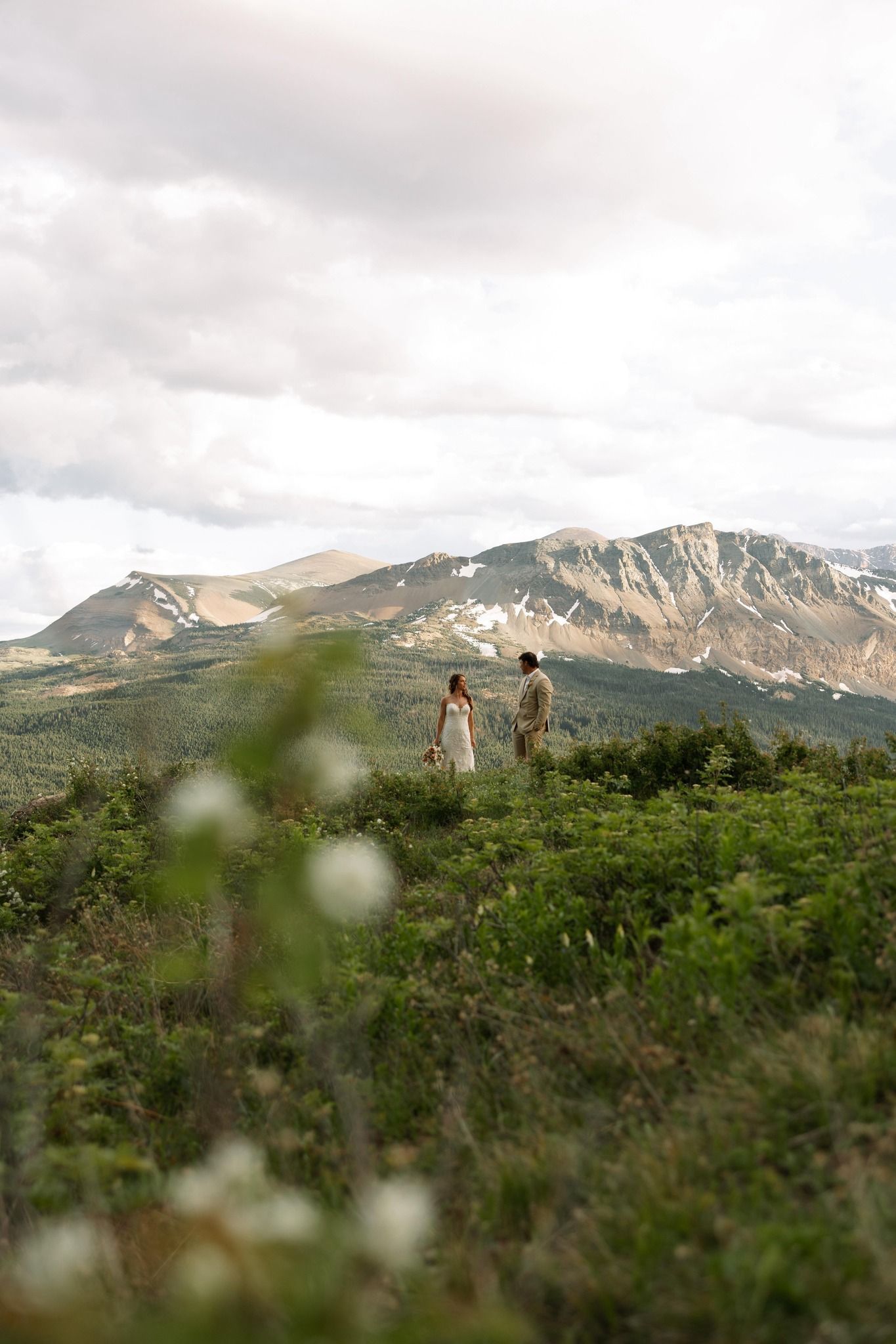 Couple stands on a hilltop, overlooking mountains. Green foliage in the foreground, cloudy sky.