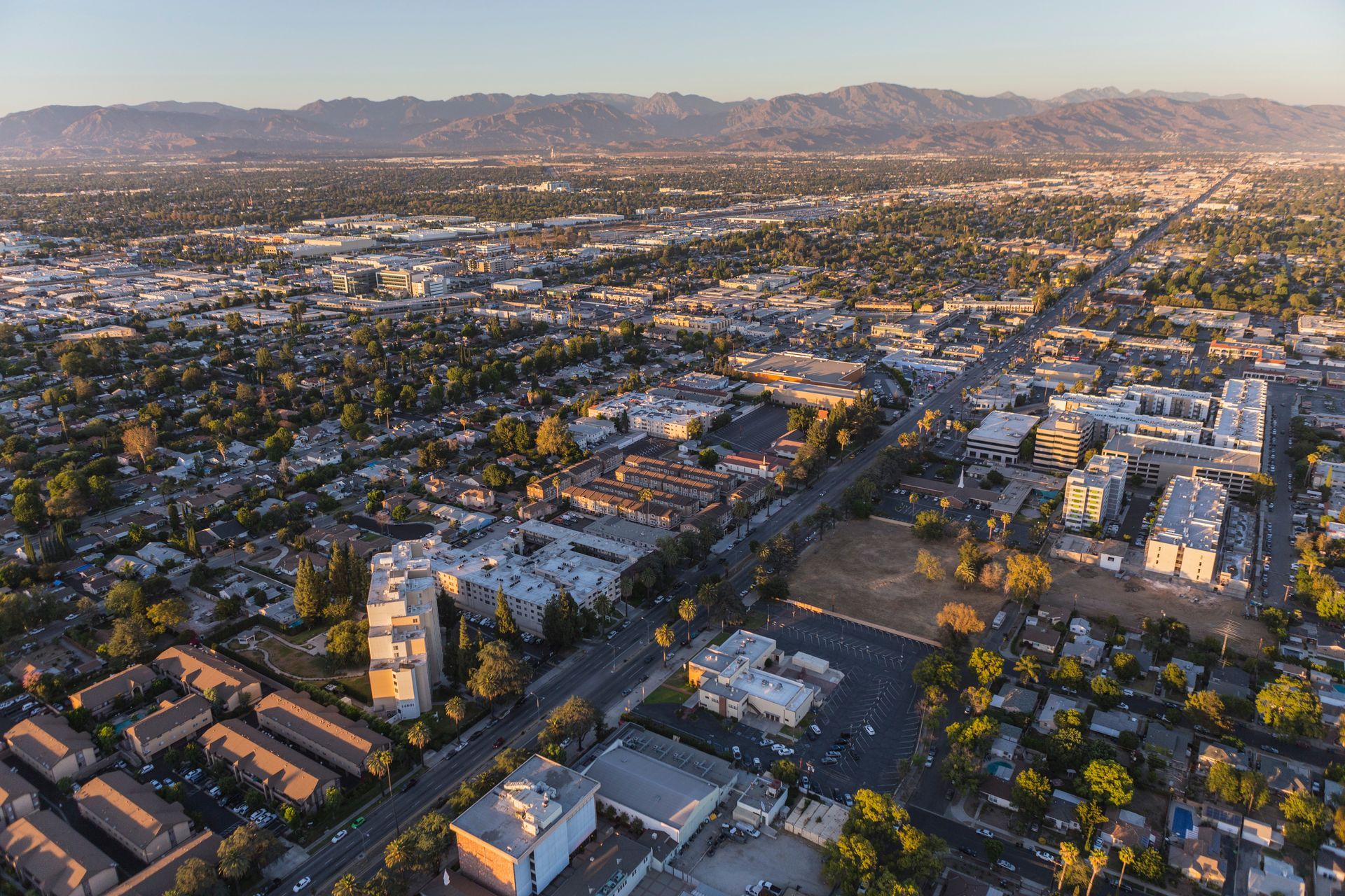 Aerial view of a city with roads, buildings, and trees under a hazy sky, mountains in the distance.