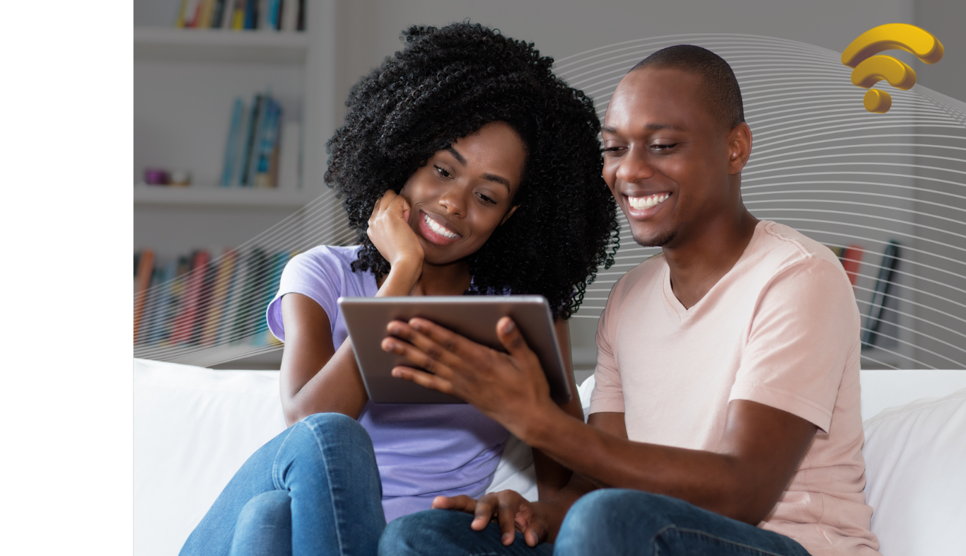 Couple smiling, looking at tablet on a couch. Living room setting, shelves in background, WiFi signal.