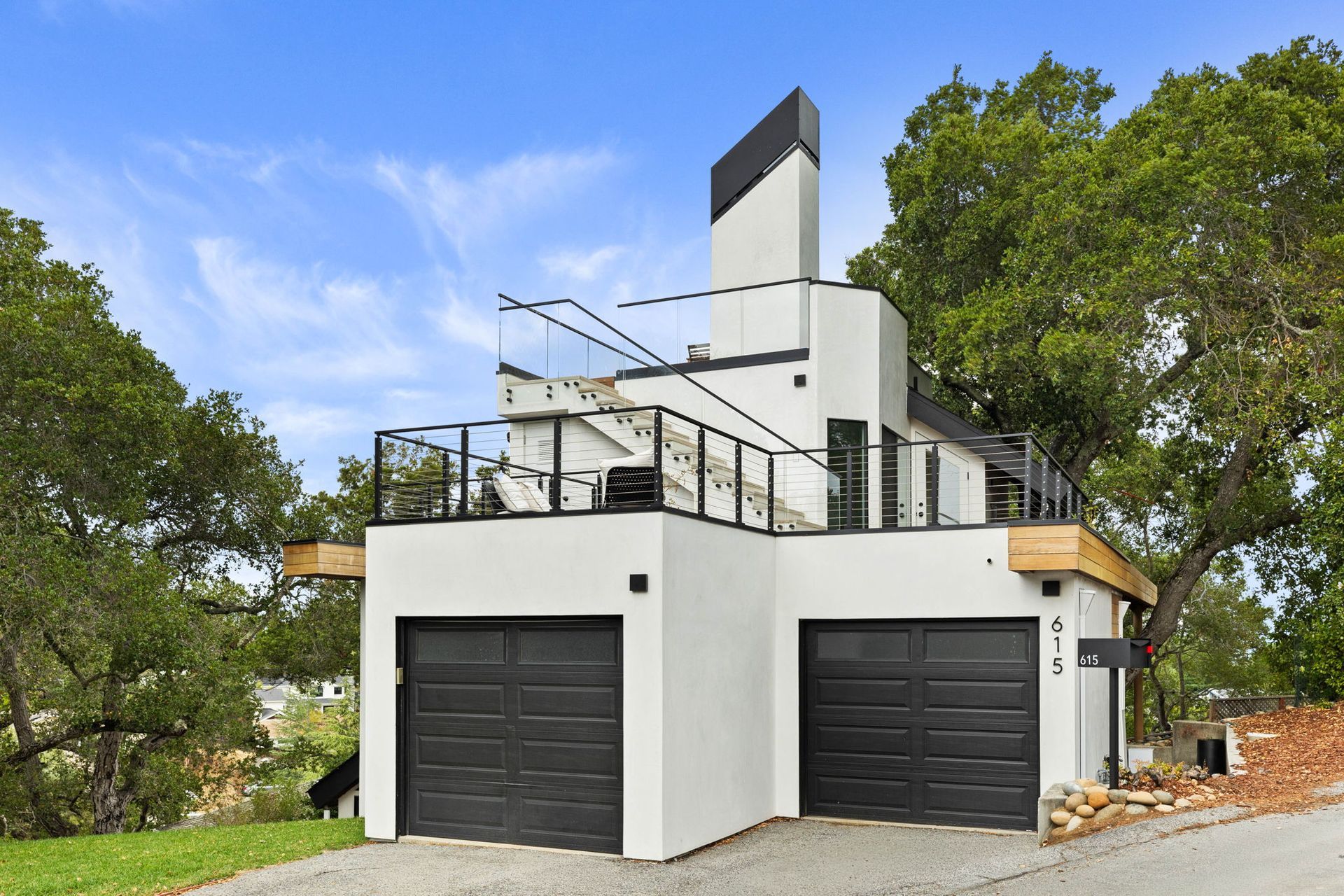 Modern white house with black garage doors, deck, and tall chimney-like structure, set amongst trees.