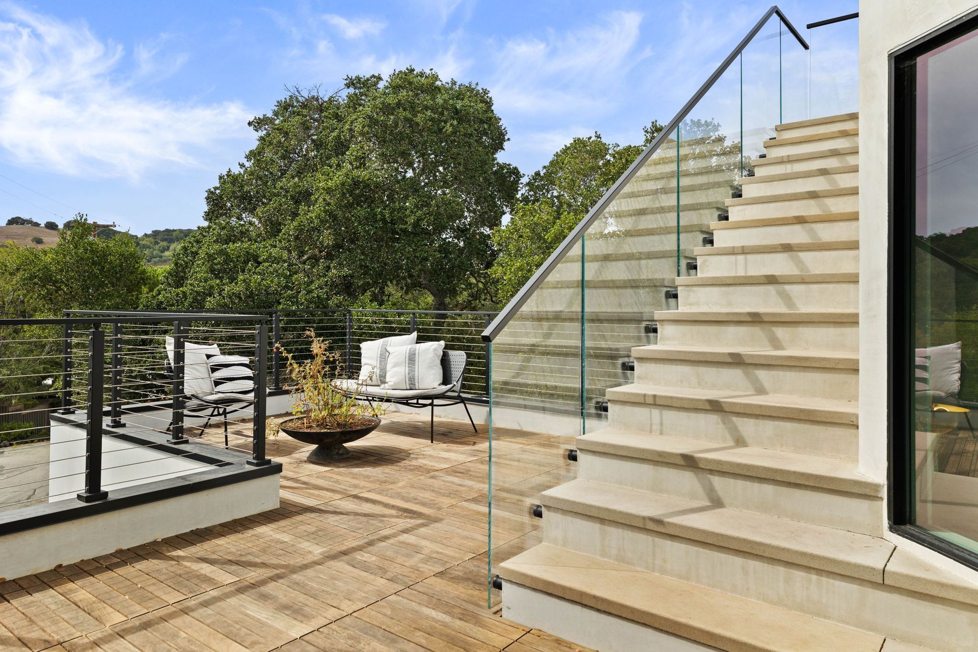Wooden deck with stairs, glass railings, and seating overlooking lush greenery under a blue sky.