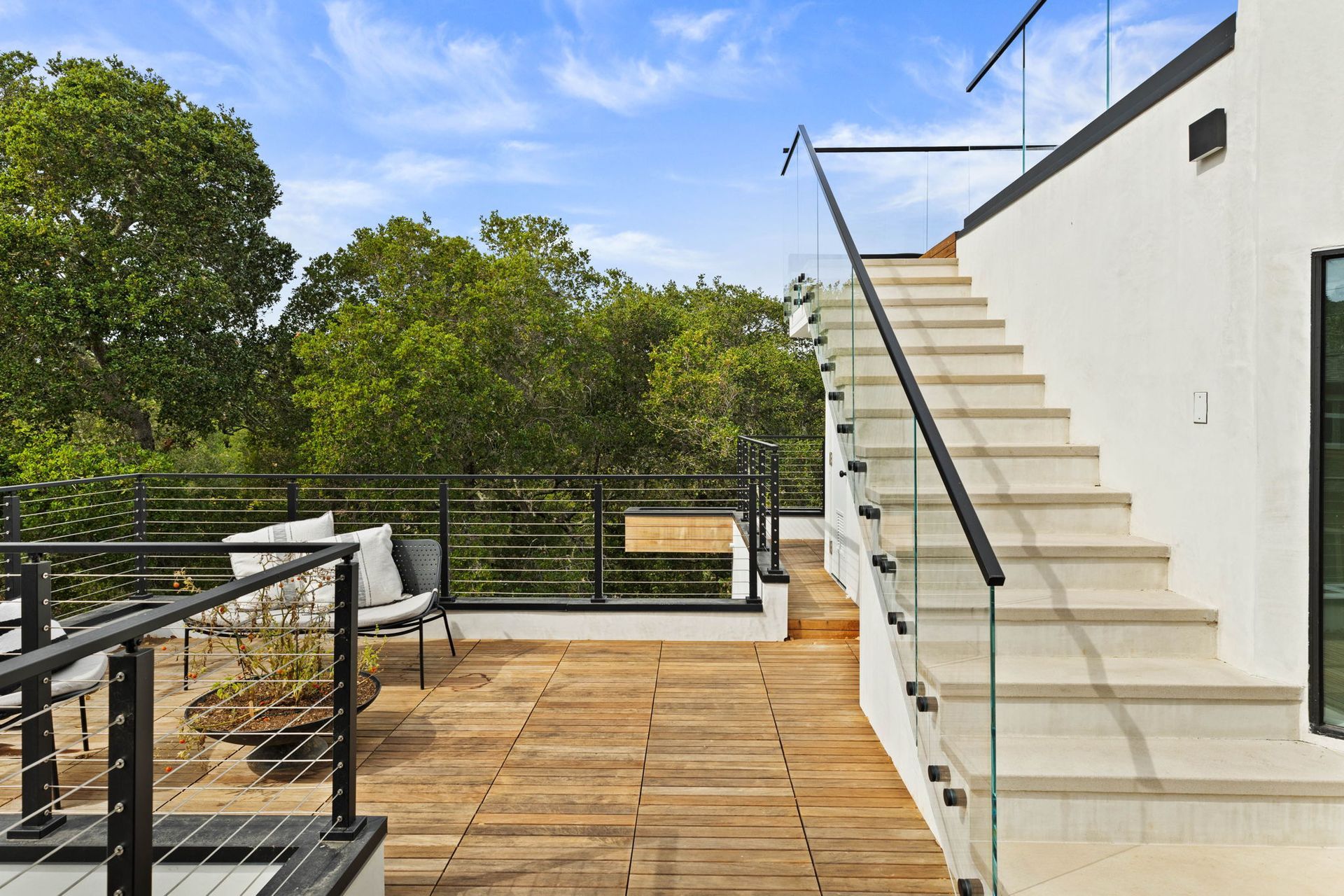 Rooftop deck with wooden flooring, stairs, and glass railings; overlooking trees under a blue sky.