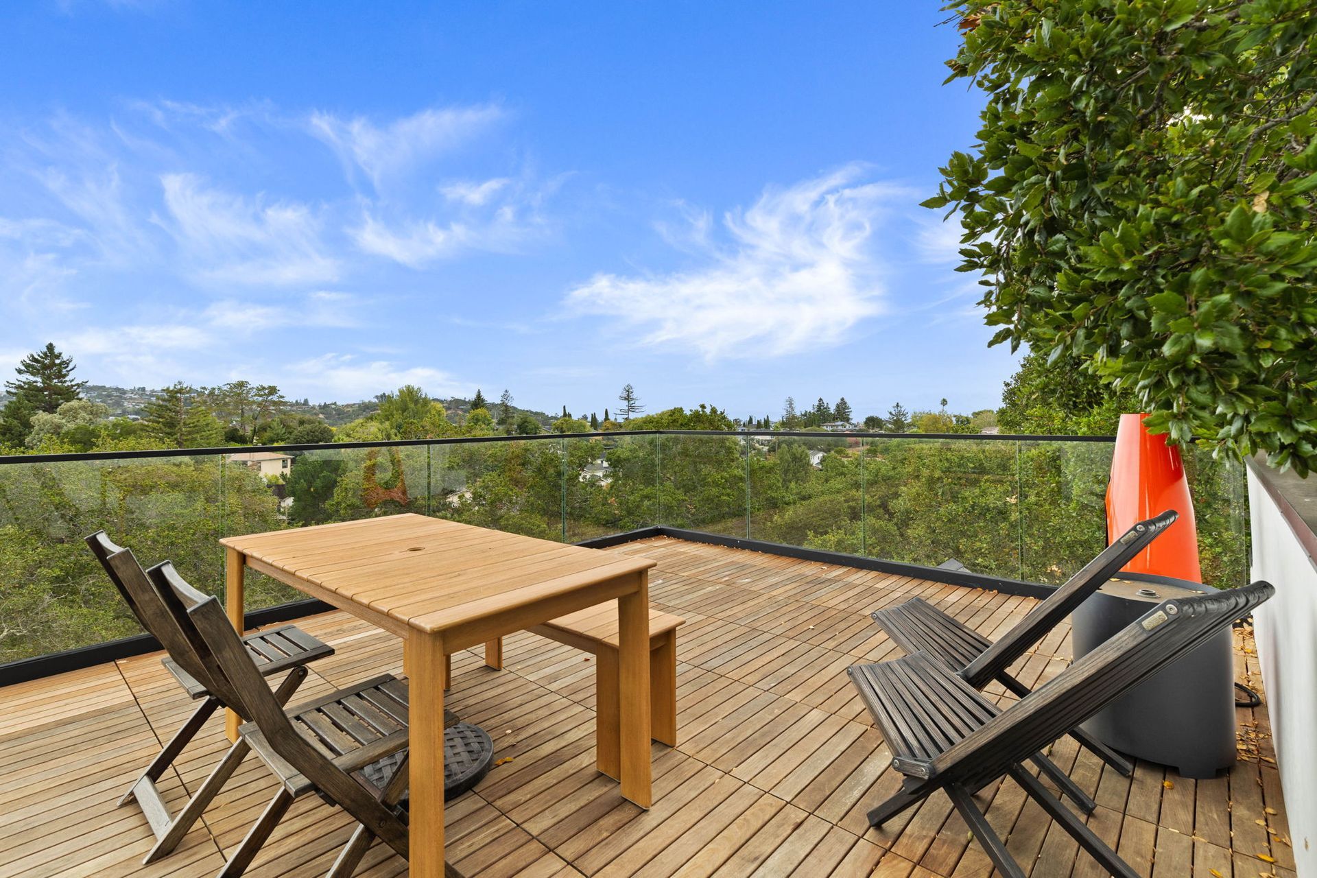 Rooftop deck with wooden table and chairs, overlooking a tree-filled landscape under a blue sky.