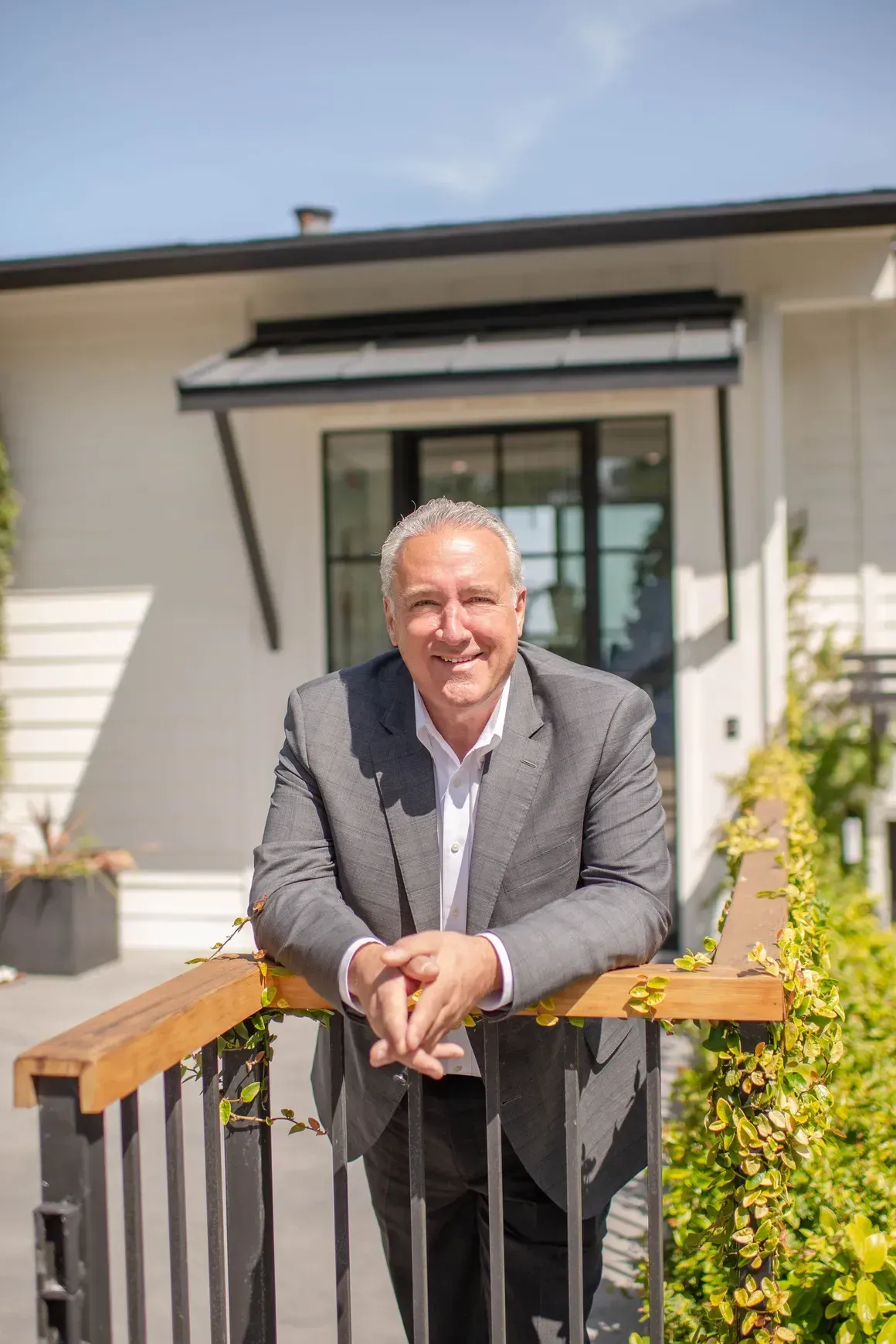 Man in a gray suit leaning on a wooden railing, smiling in front of a modern white building.