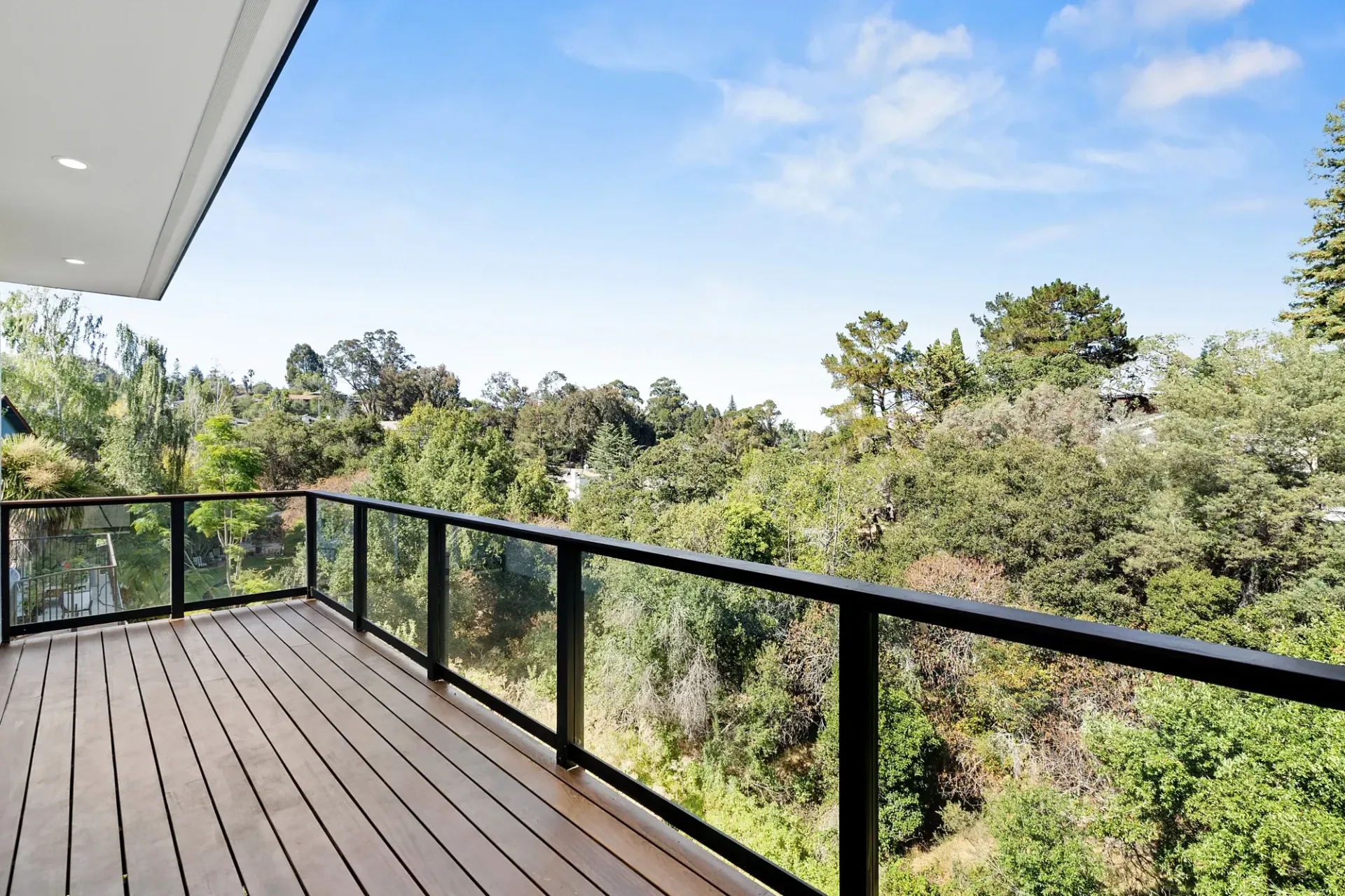 Wooden deck with glass railing overlooks lush green trees under a blue sky.