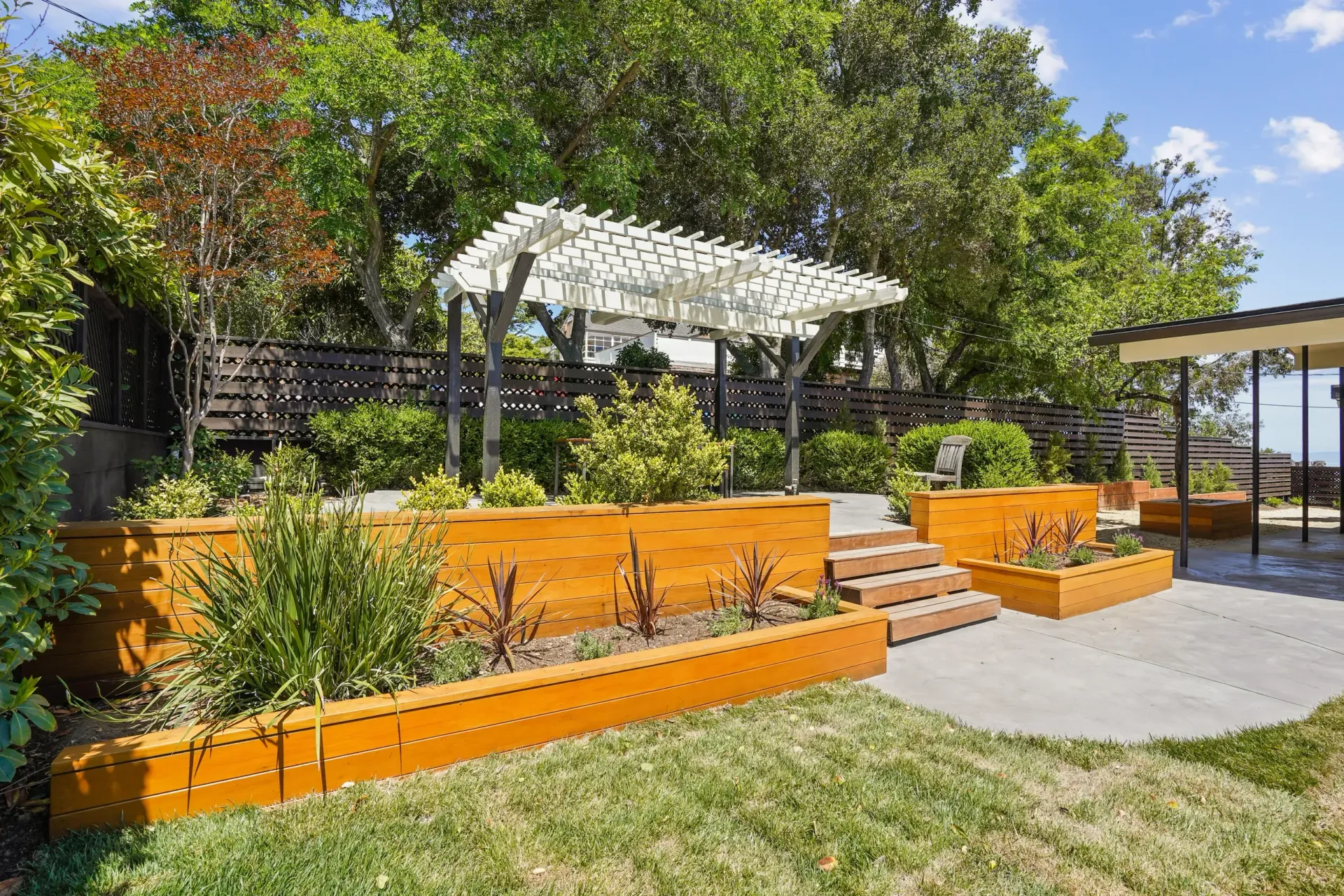 Backyard with tiered orange planters, pergola, steps, and trees against a sunny sky.