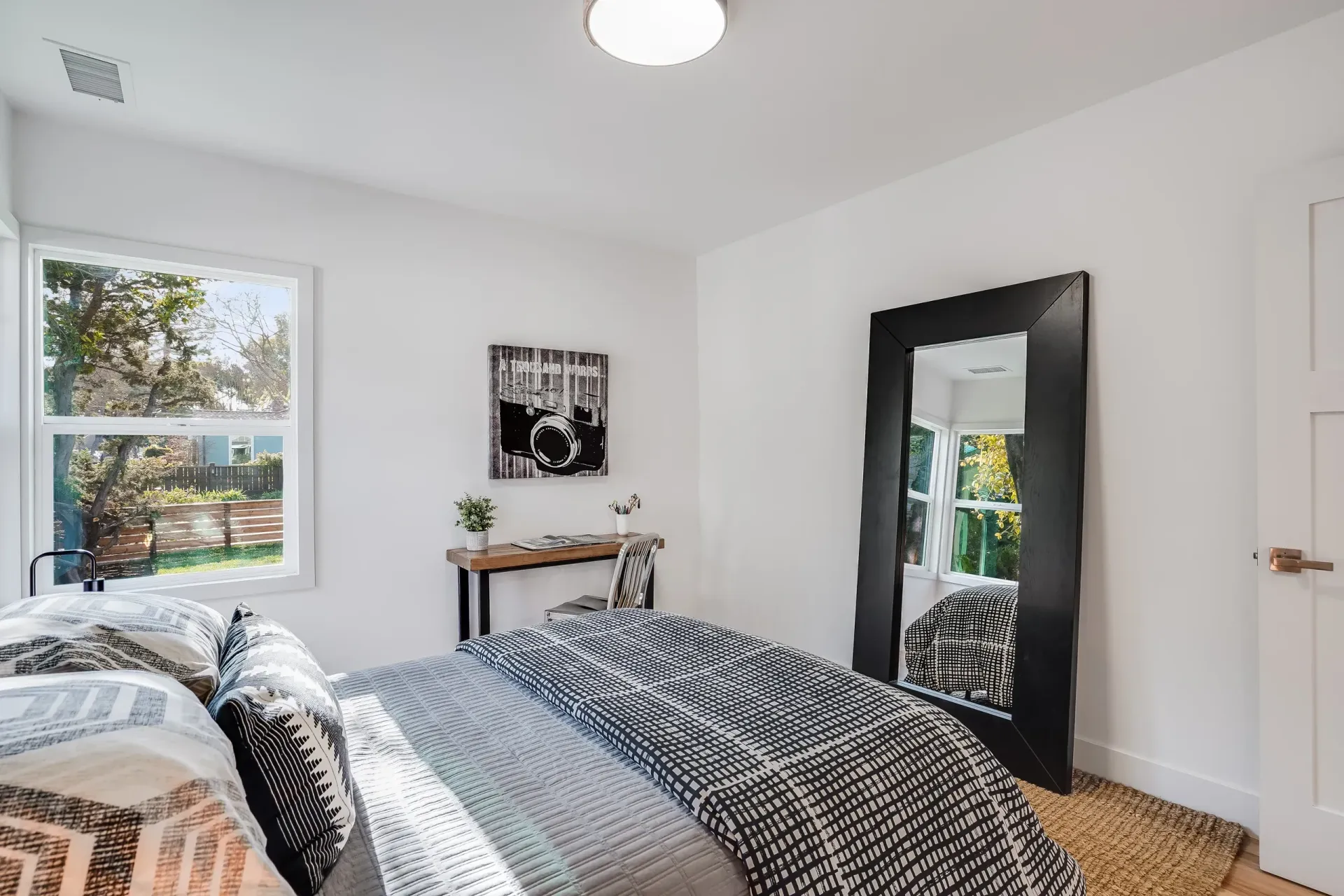 Bedroom with bed, large mirror, small desk, and window overlooking a yard. White walls, natural light.