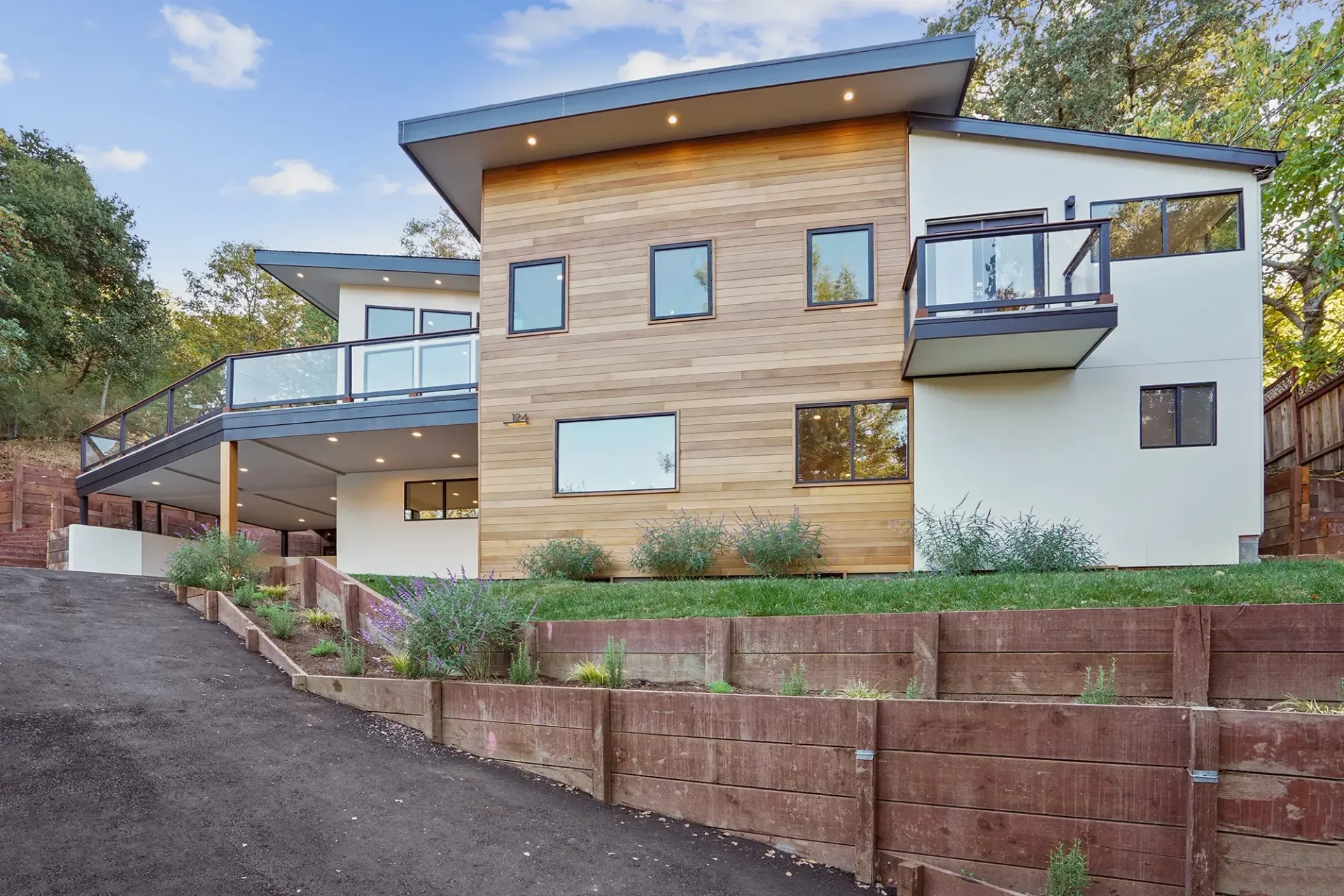 Modern two-story house with wood siding, balconies, and a sloped roof, set in a hillside landscape.