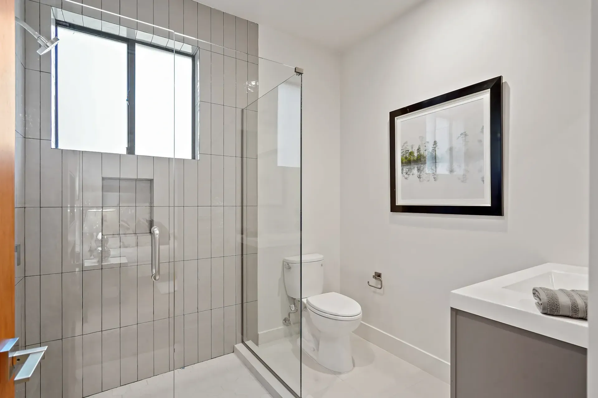 Modern bathroom with shower, toilet, and vanity. Gray tile wall, glass shower door. Art on the wall.