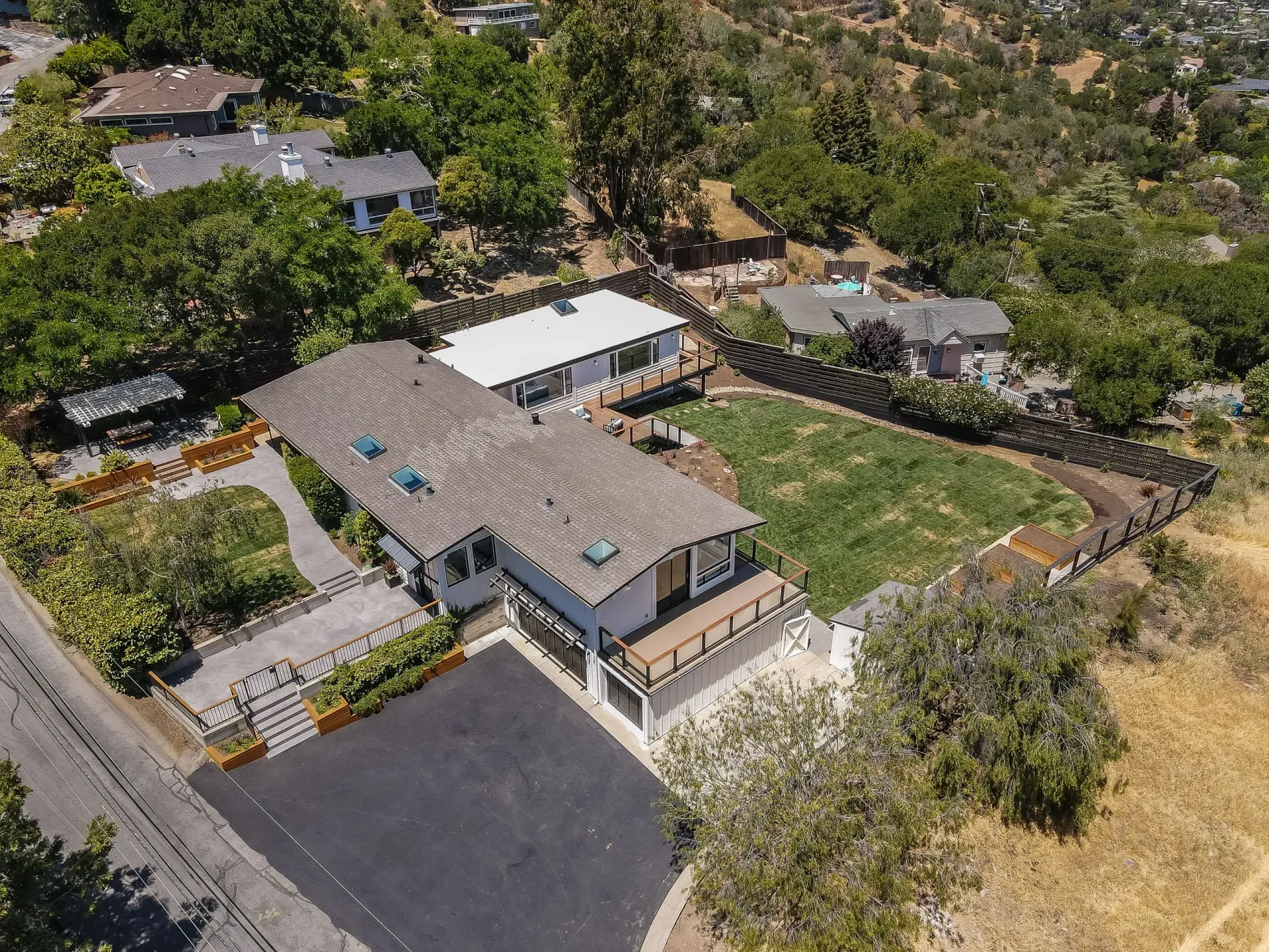 Aerial view of homes nestled in hillside greenery; asphalt driveway, fenced yards.