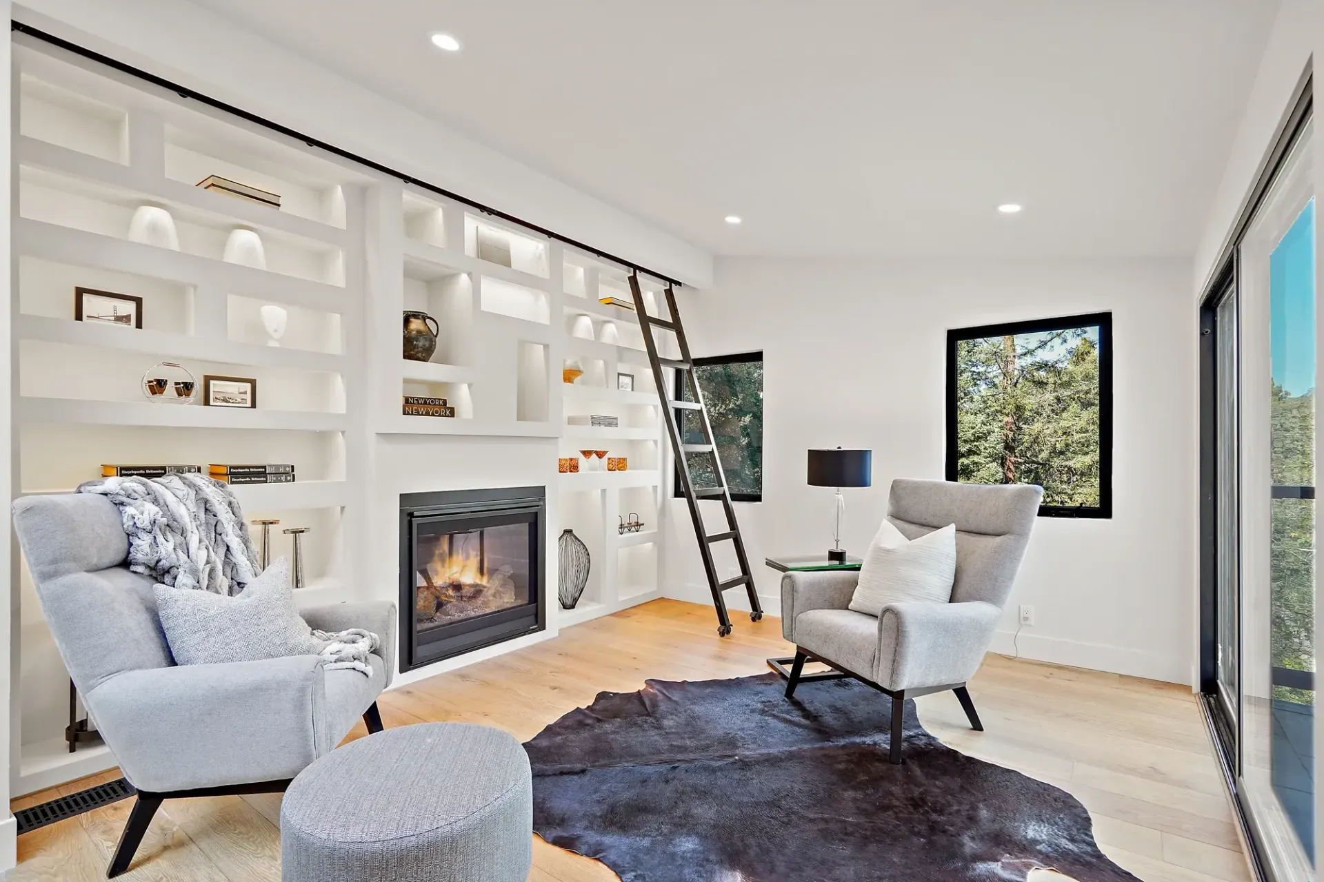 Cozy living room with fireplace, built-in shelves, two armchairs, a cowhide rug, and a rolling ladder.