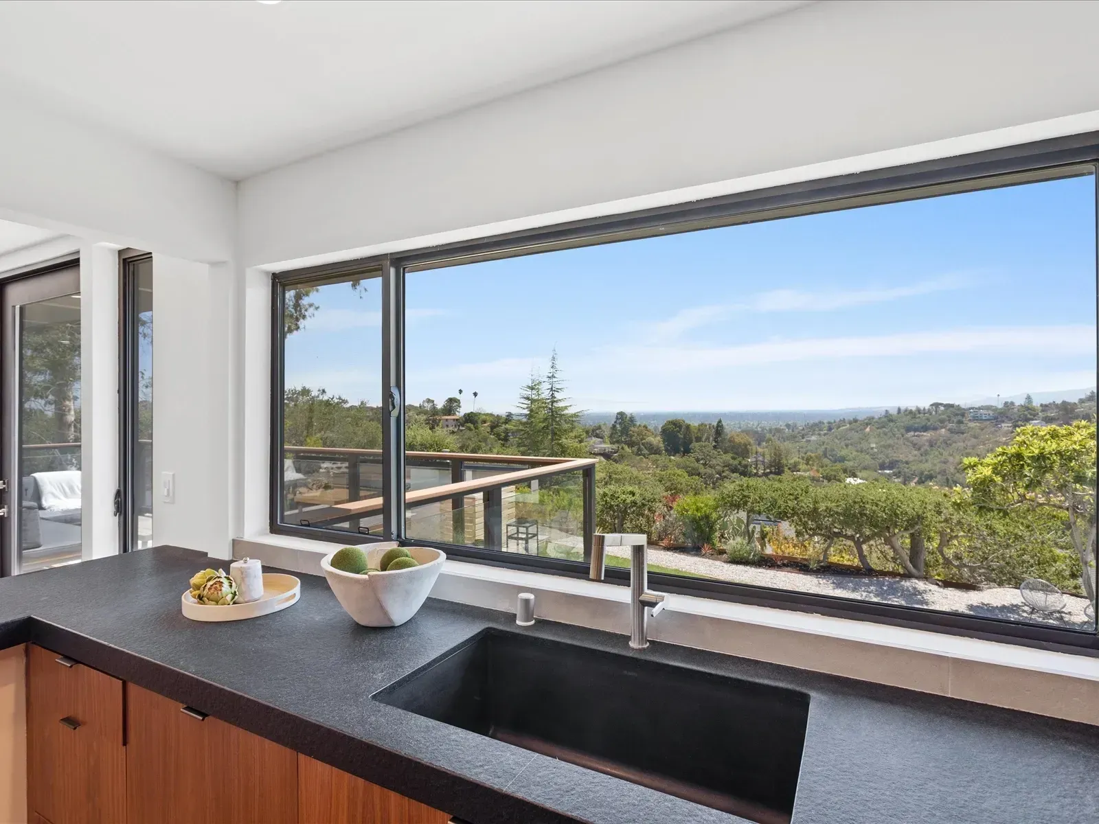 Kitchen with large window overlooking a scenic valley. Black countertop, dark sink, light wooden cabinets.