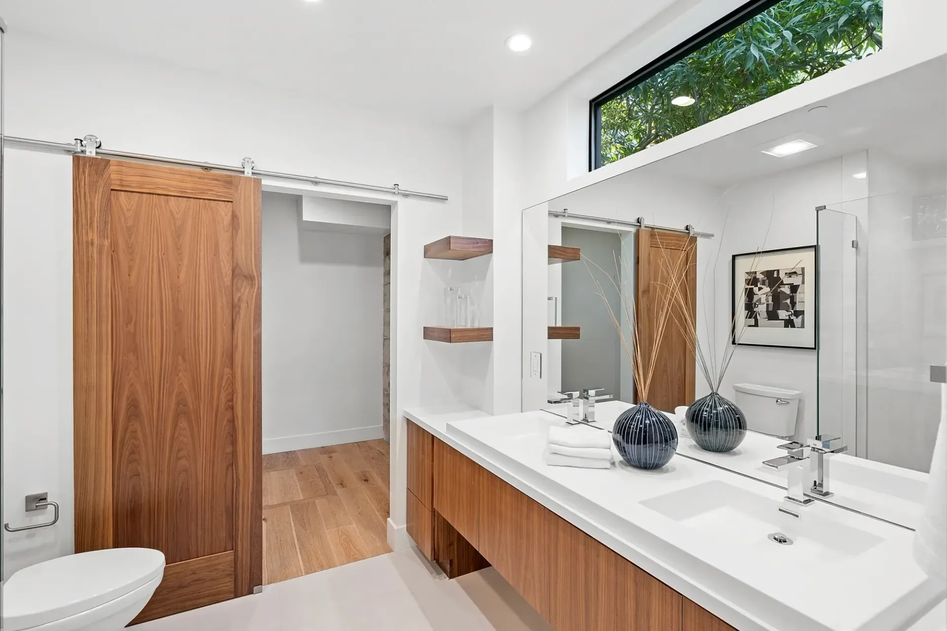 Bathroom with wooden vanity, sliding door, large mirror, and window with greenery view.