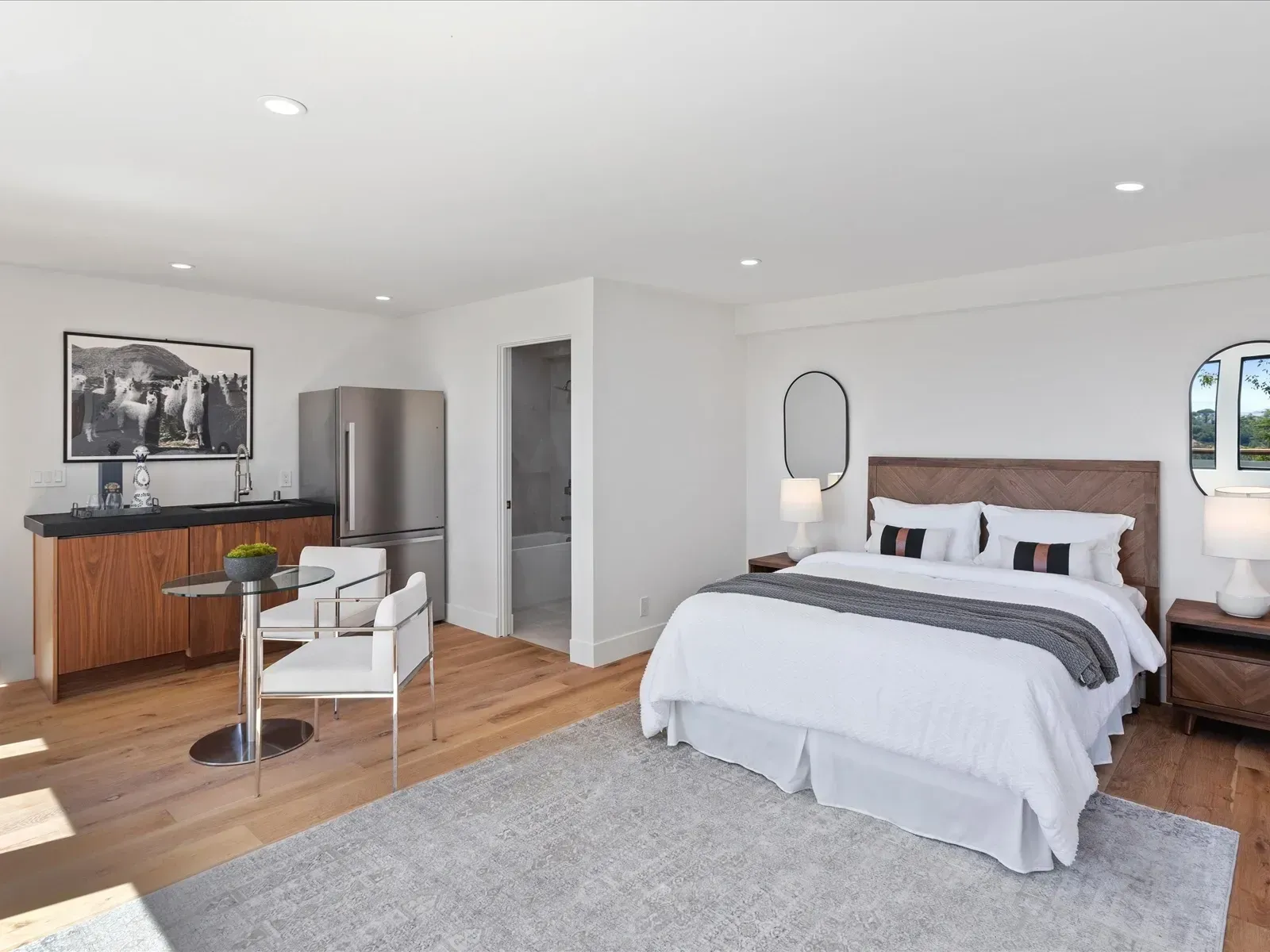 Bedroom studio with wooden floor, bed, kitchenette, and round table. Bright and minimalist.