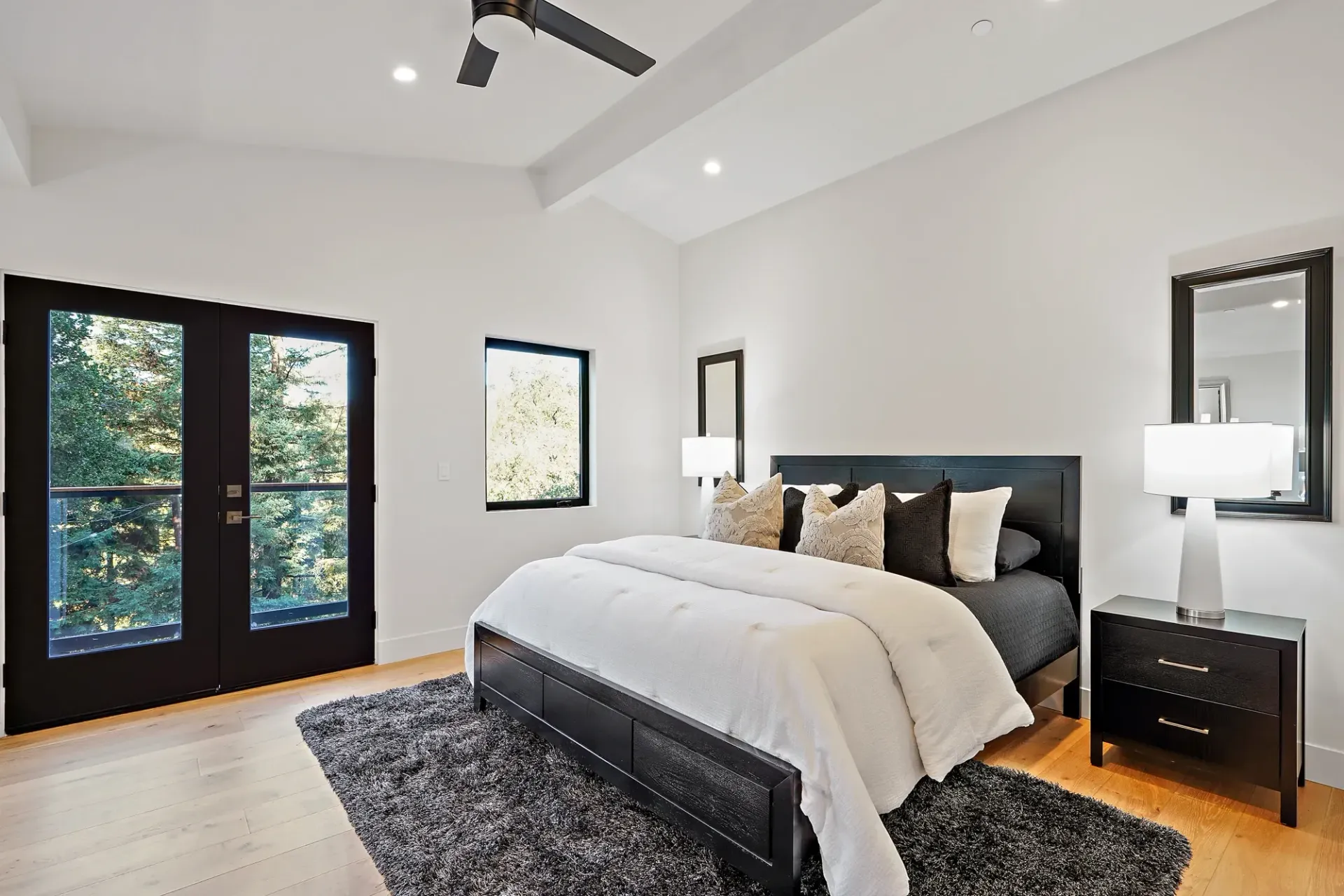 Bedroom with black accents: bed, doors, windows, rug, lamps, mirrors. White walls, wood floor, ceiling fan.